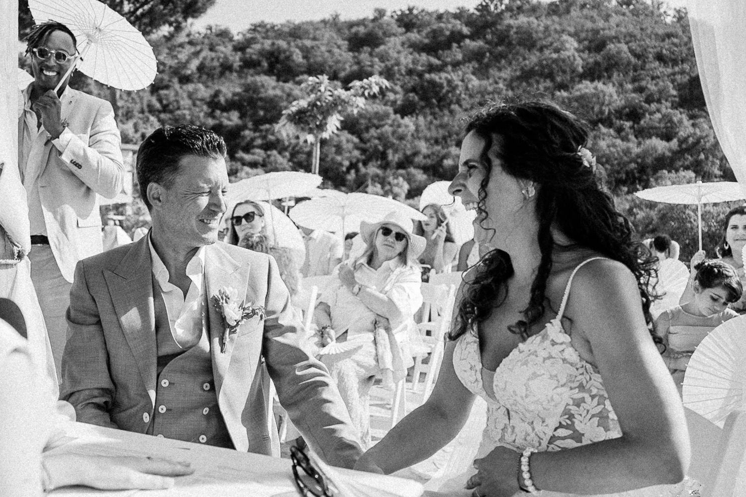 Black and white photo of a smiling bride and groom exchanging vows at an outdoor wedding ceremony with guests.