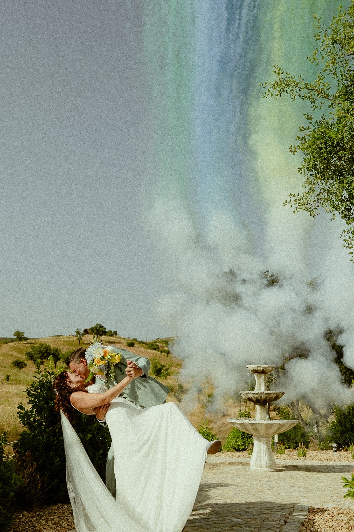 Bride and groom embrace with colorful rainbow smoke bomb at outdoor wedding near stone fountain.