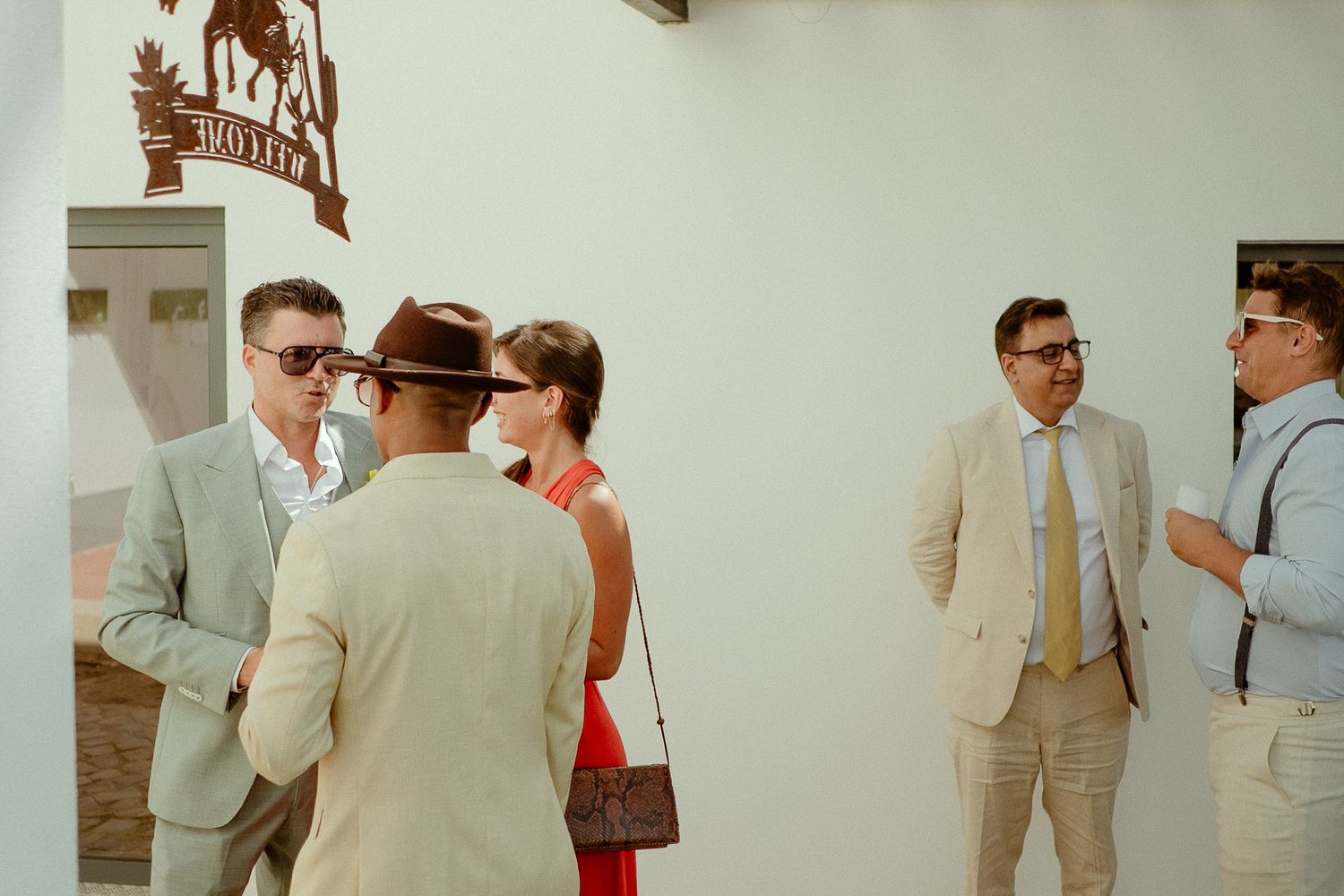 Elegantly dressed guests in suits and sunglasses socializing outside a white building at a summer wedding event.