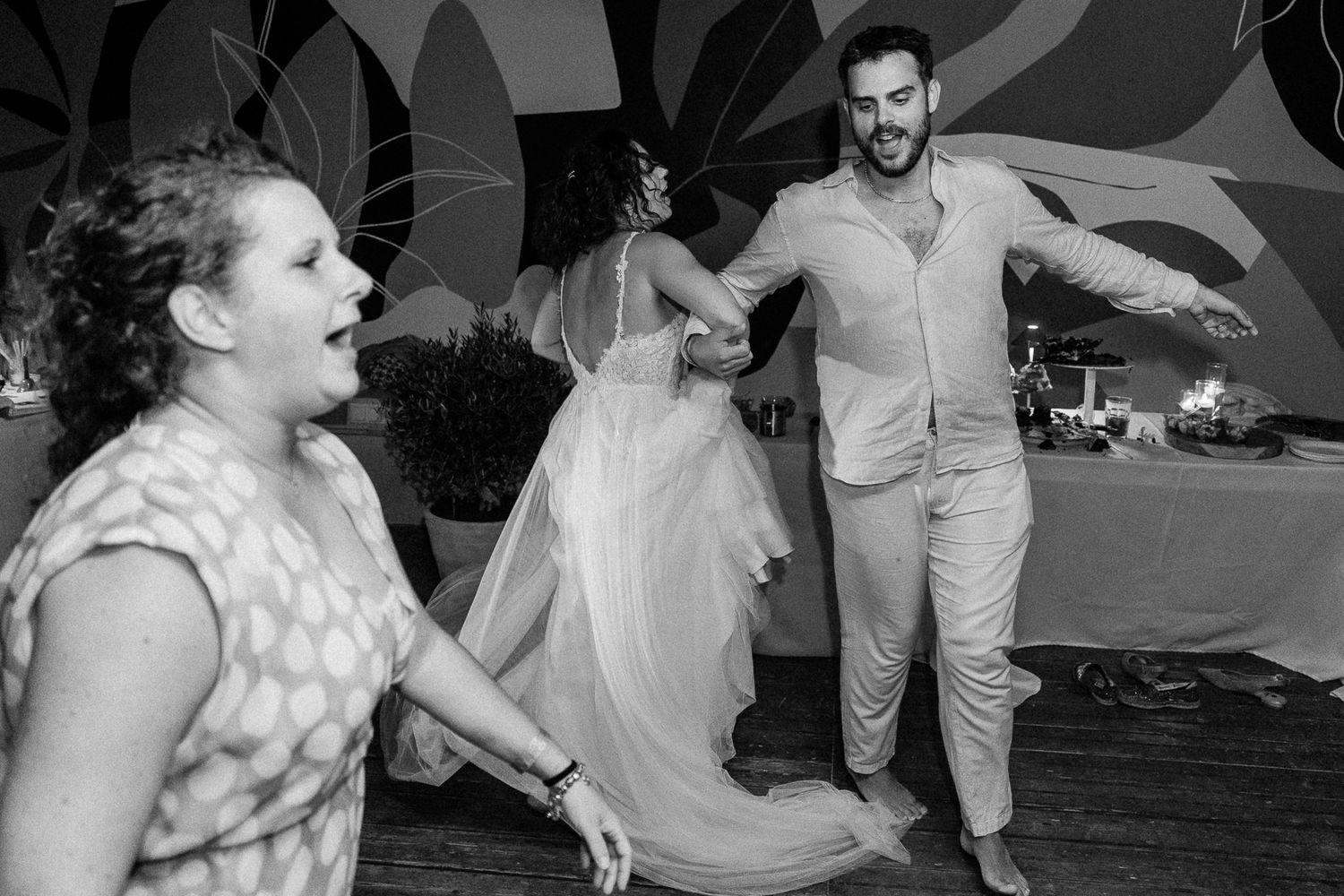 Black and white photo of bride and groom dancing joyfully with a guest at their wedding reception.
