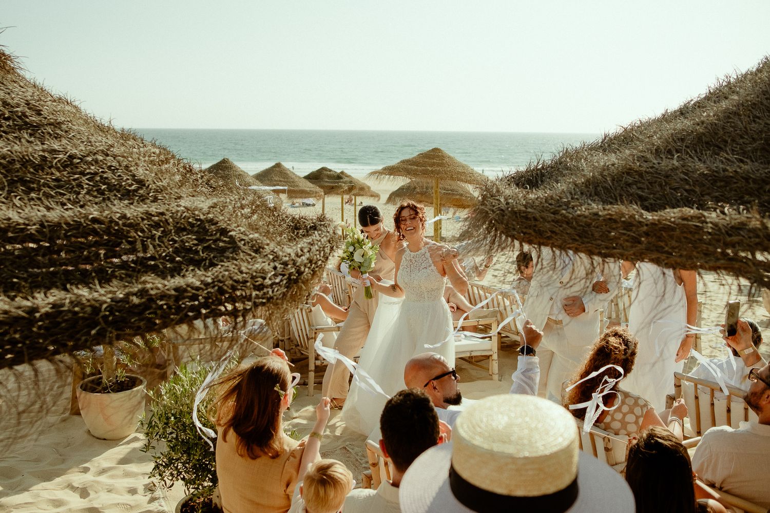 Bride and groom celebrate at a beachside wedding ceremony with thatched umbrellas and ocean views.