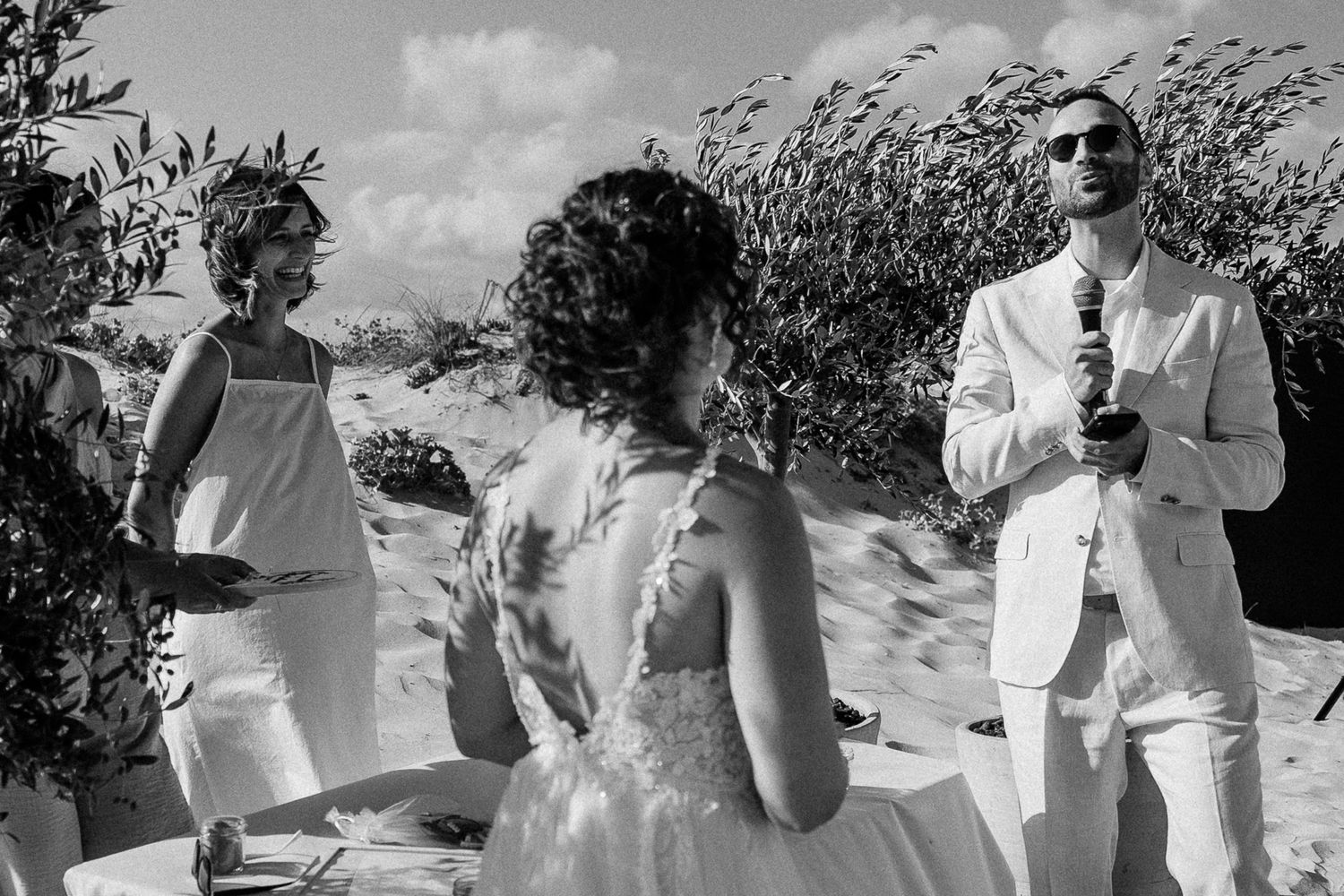 Black and white beach wedding ceremony with bride, groom in white suit, and officiant among sand dunes.