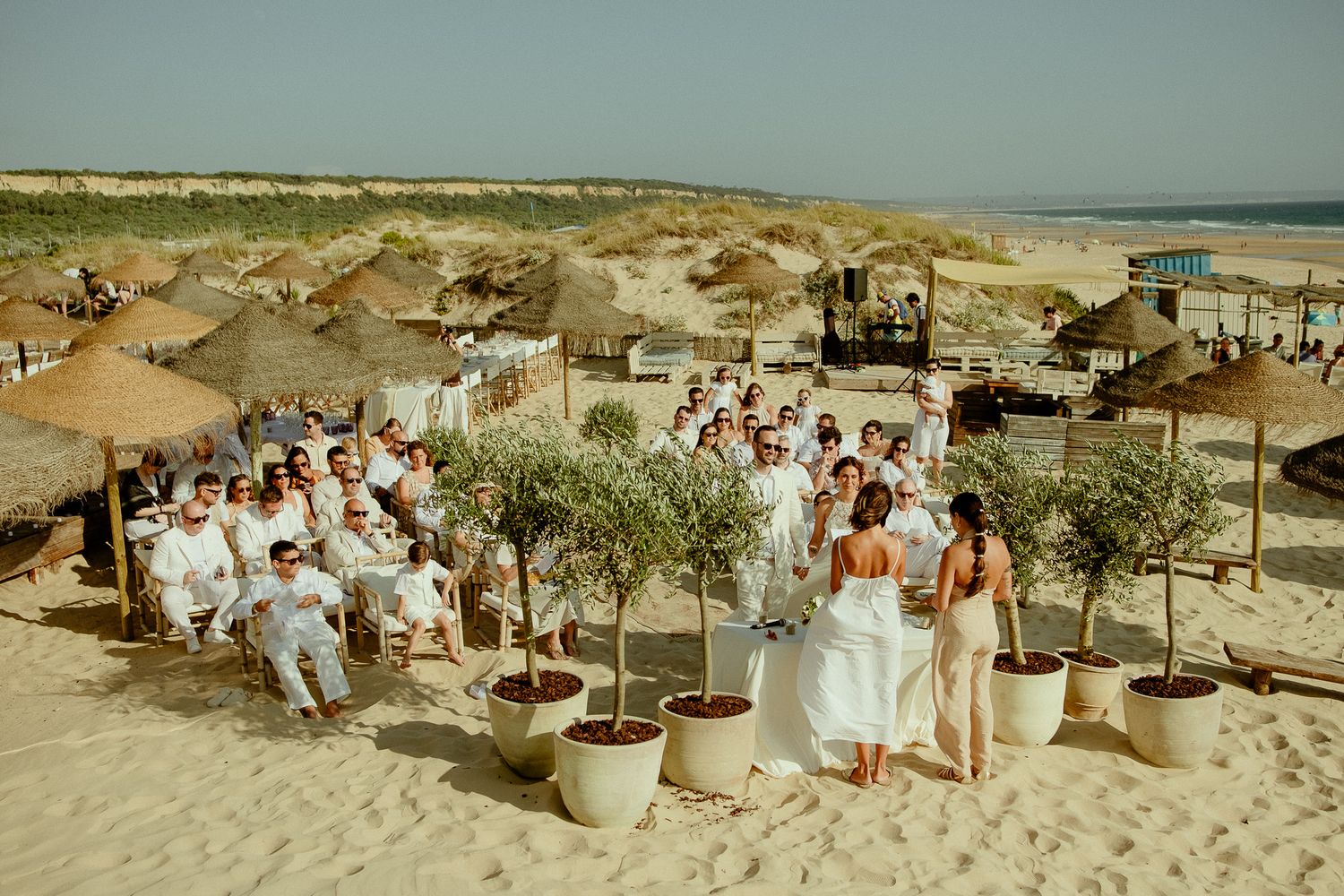 Outdoor beach wedding ceremony with guests in white attire, potted olive trees, and straw umbrellas on sandy shore.