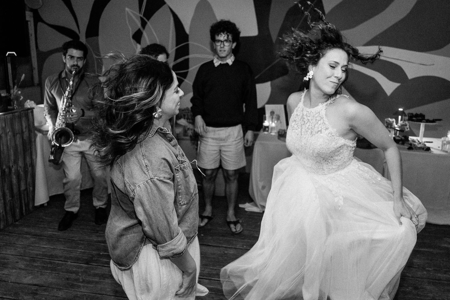 Two women dancing joyfully at a wedding reception in black and white photo with live band performing in background.