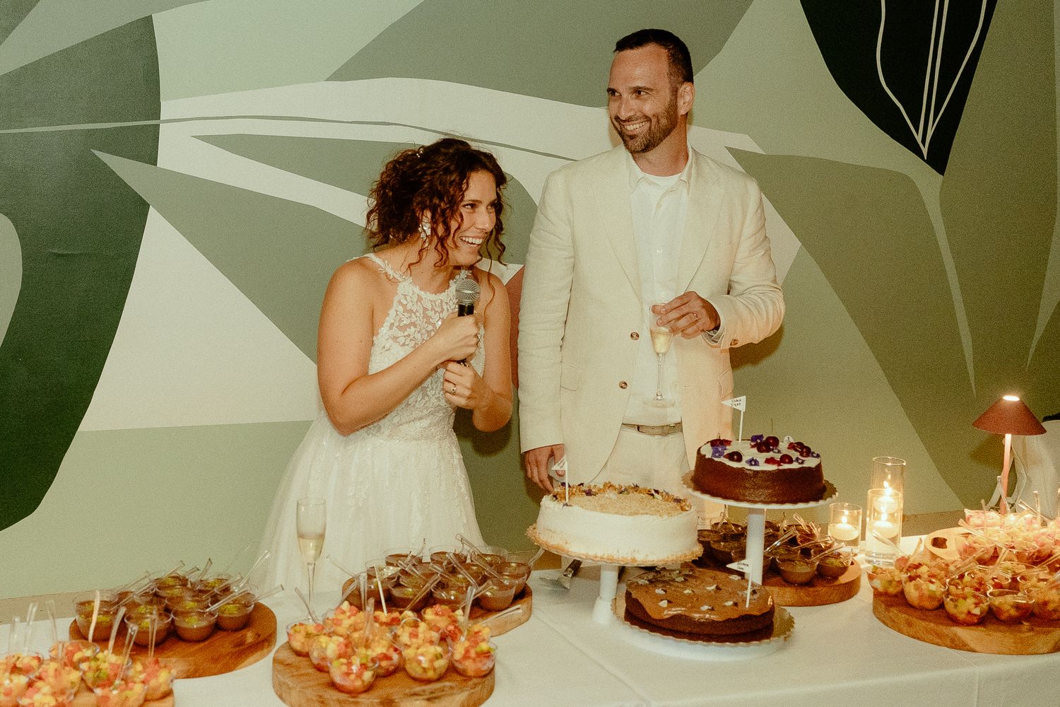 Bride and groom cutting wedding cake at dessert table with candles and pastries, standing before green botanical wall.