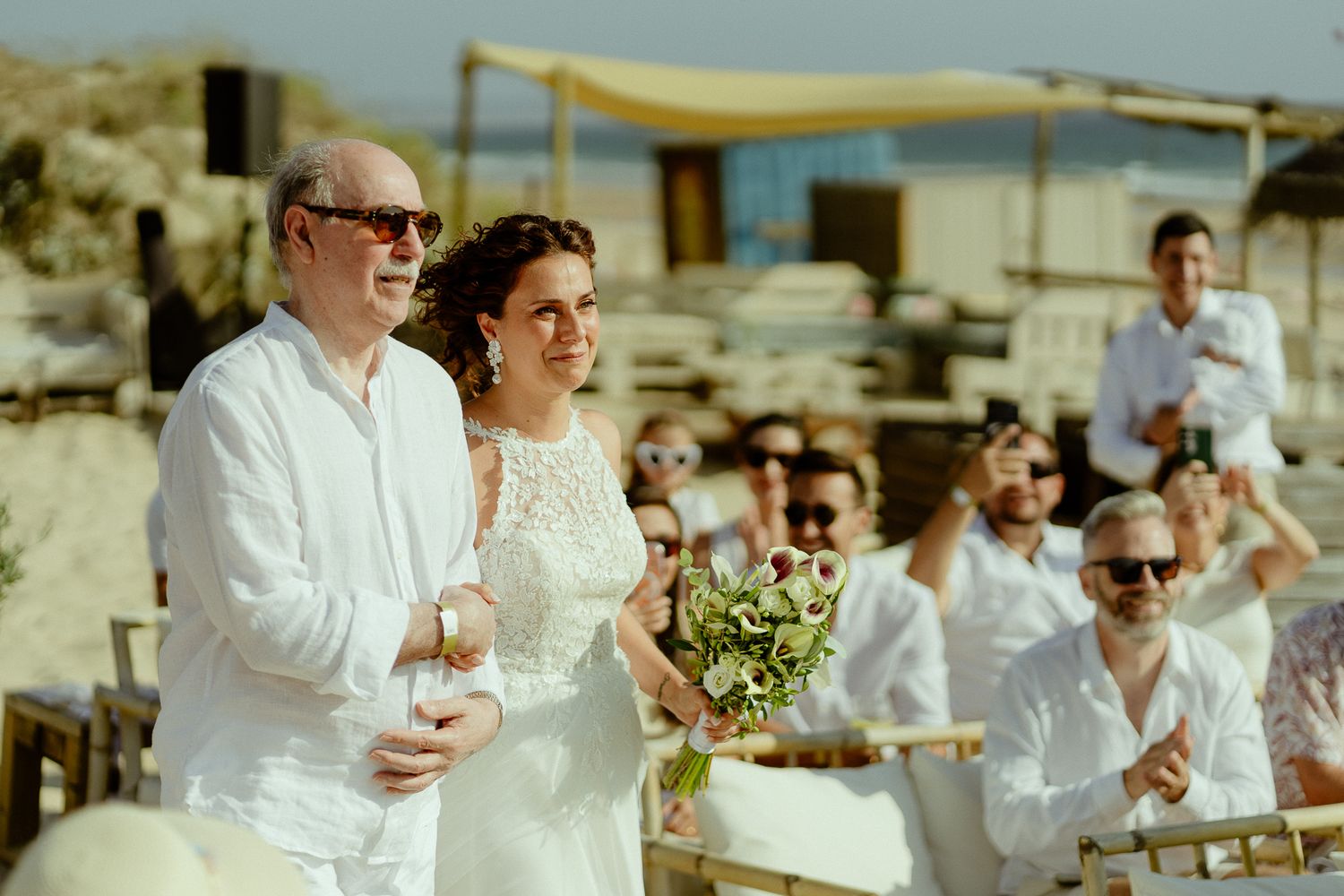 Bride and groom in white attire smile during outdoor beach wedding ceremony with guests applauding in background.