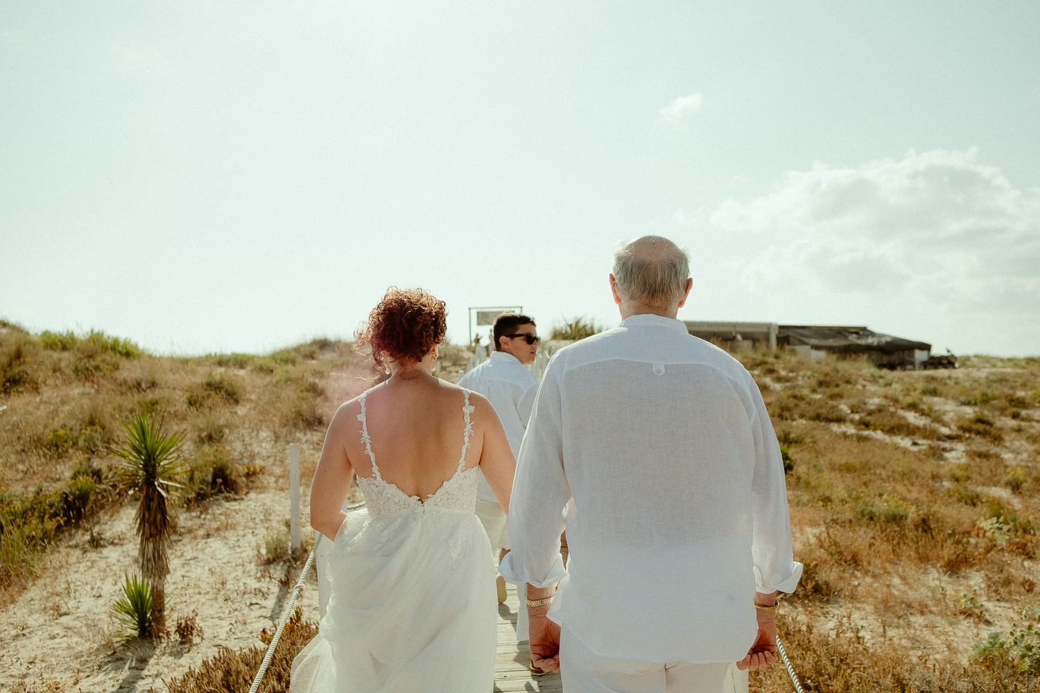 Couple in white attire walking hand-in-hand on a sandy beach path with dunes and a sunny sky.