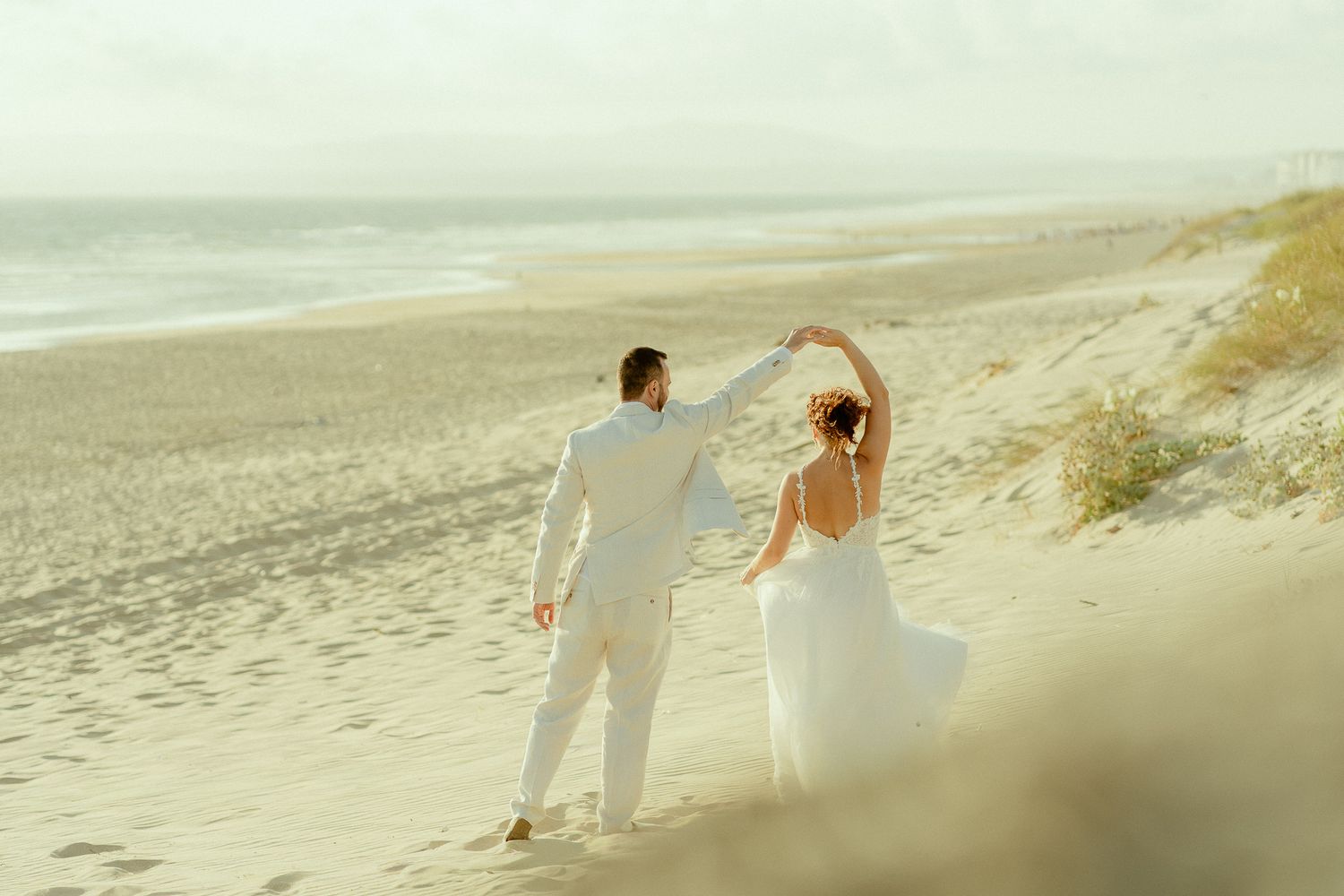 Bride and groom dancing on a sandy beach, wearing white wedding attire with ocean waves in the background.
