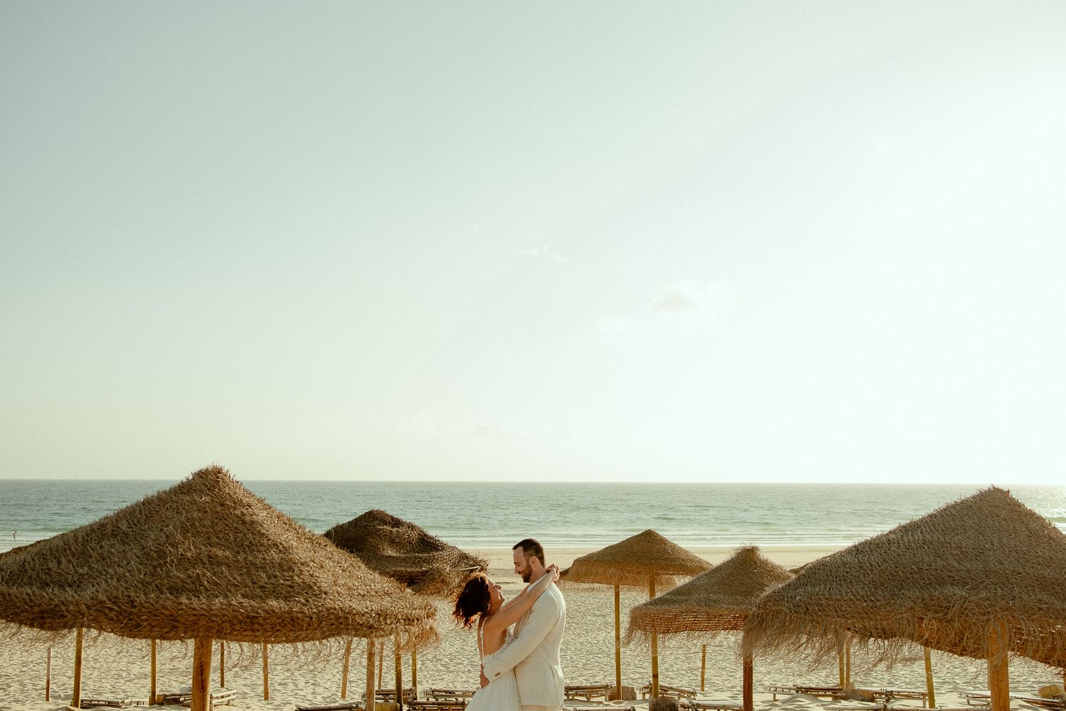 Couple embracing on a beach with thatched umbrellas and ocean backdrop on a sunny day.