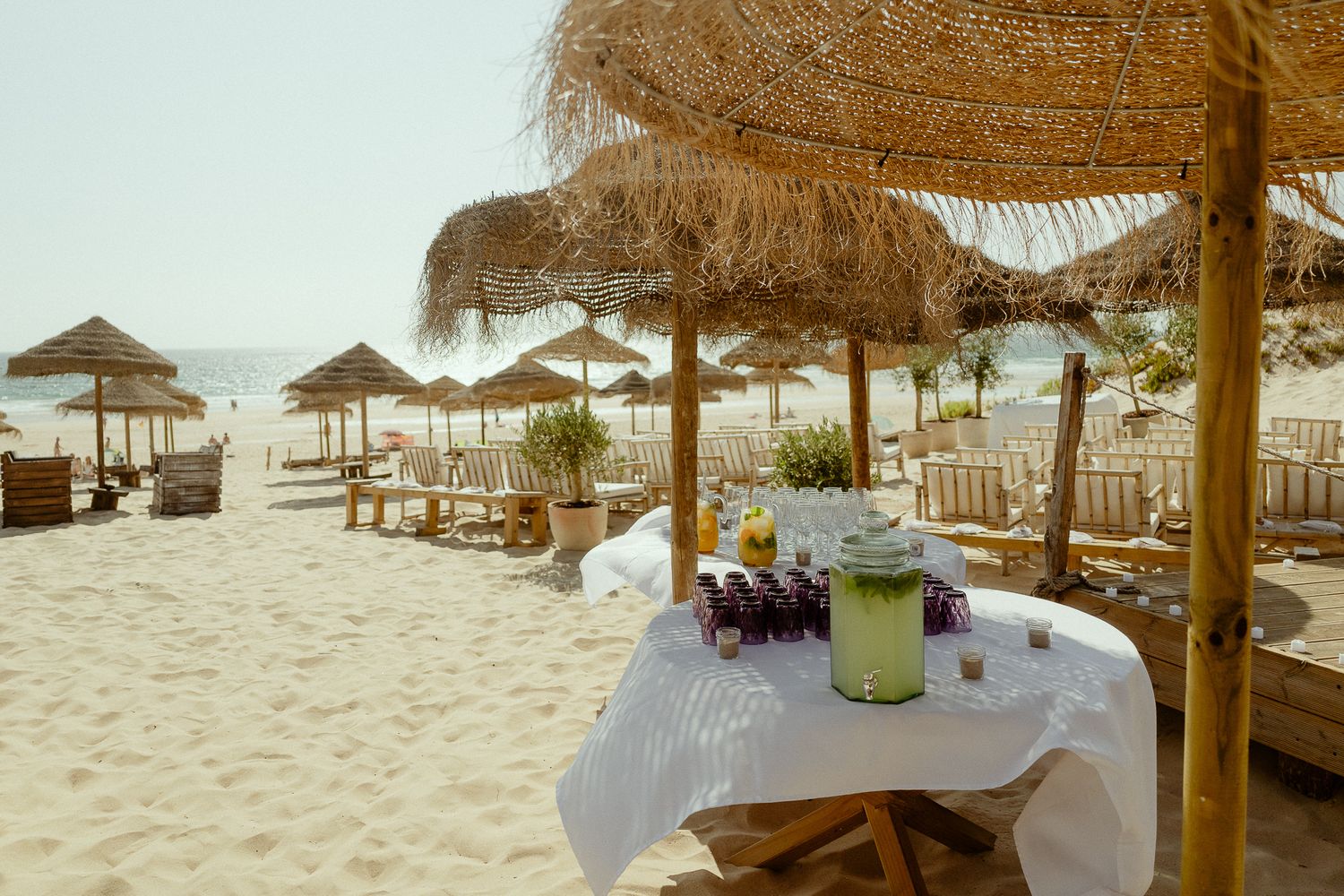Sandy beach club with thatched umbrellas, white tablecloths, and tropical drinks on a sunny day.