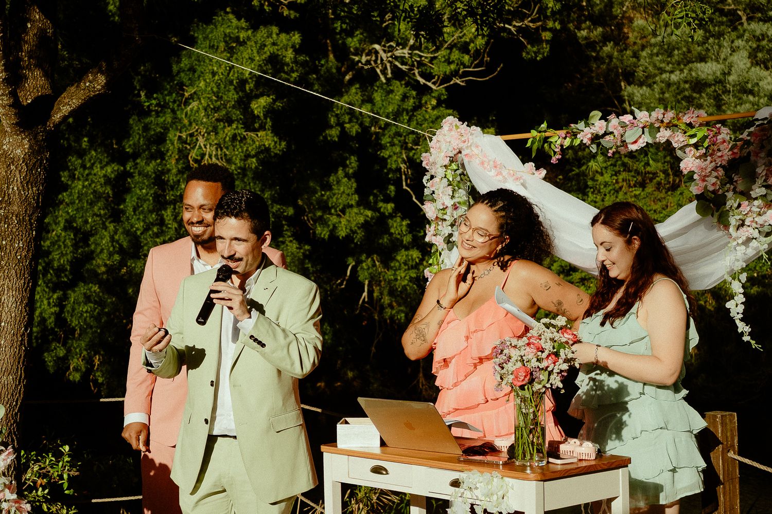 Outdoor wedding ceremony with guests laughing near floral arch, man in pink suit speaking into microphone.