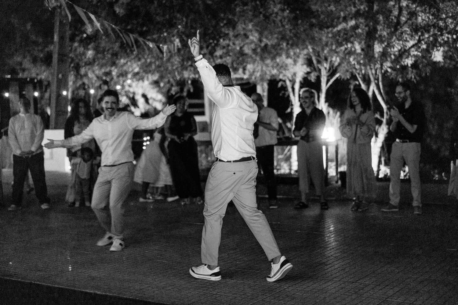 Black and white photo of people dancing outdoors at a nighttime event with trees and lights in the background.