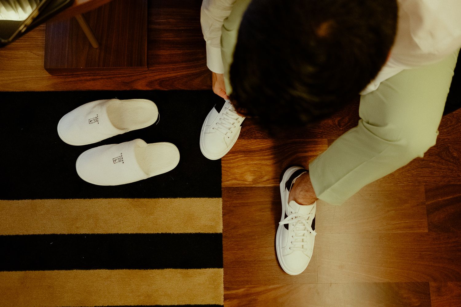 Person placing white sneakers beside hotel slippers on a striped rug near wooden flooring.