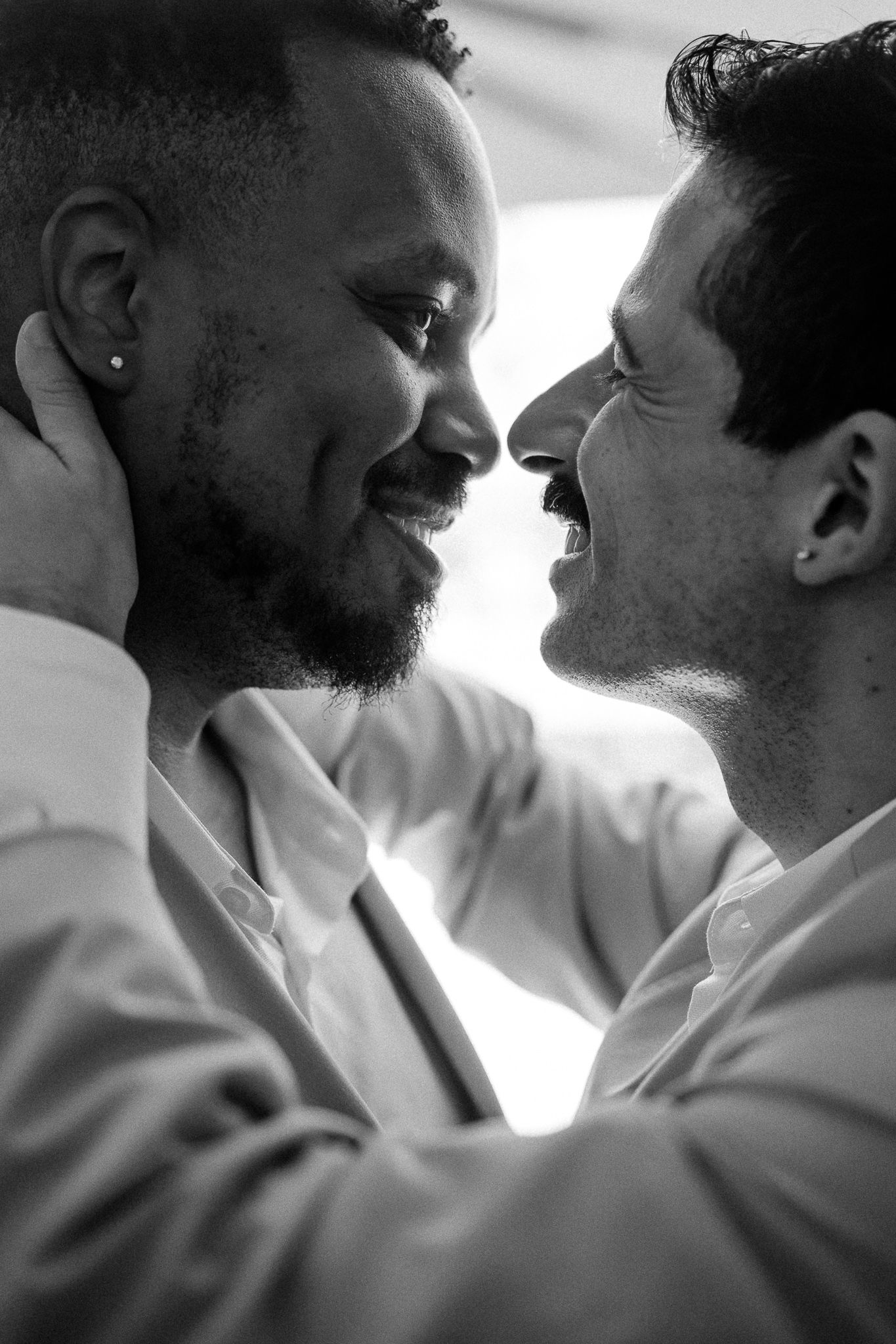 Black and white photo of two smiling men in white shirts sharing an intimate moment, facing each other closely.