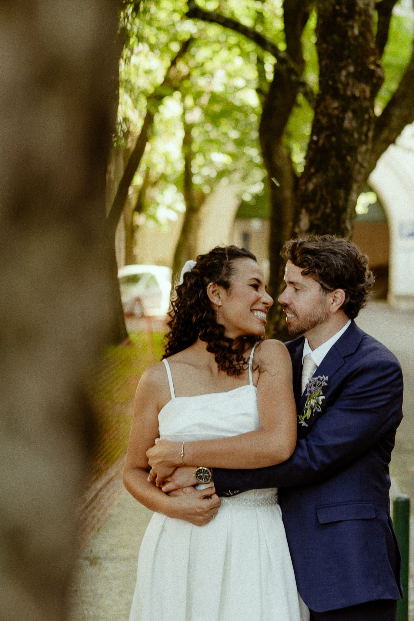 A smiling bride in white dress embraces groom in navy suit outdoors among green trees on their wedding day.