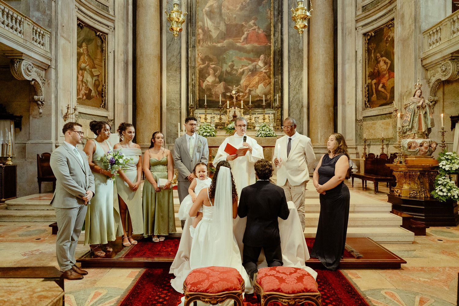 Elegant Catholic church wedding ceremony with bride and groom kneeling at altar, surrounded by wedding party in ornate baroque interior.