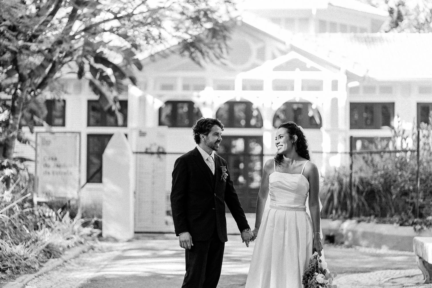 Black and white photo of a bride and groom holding hands, smiling at each other outside a white building.
