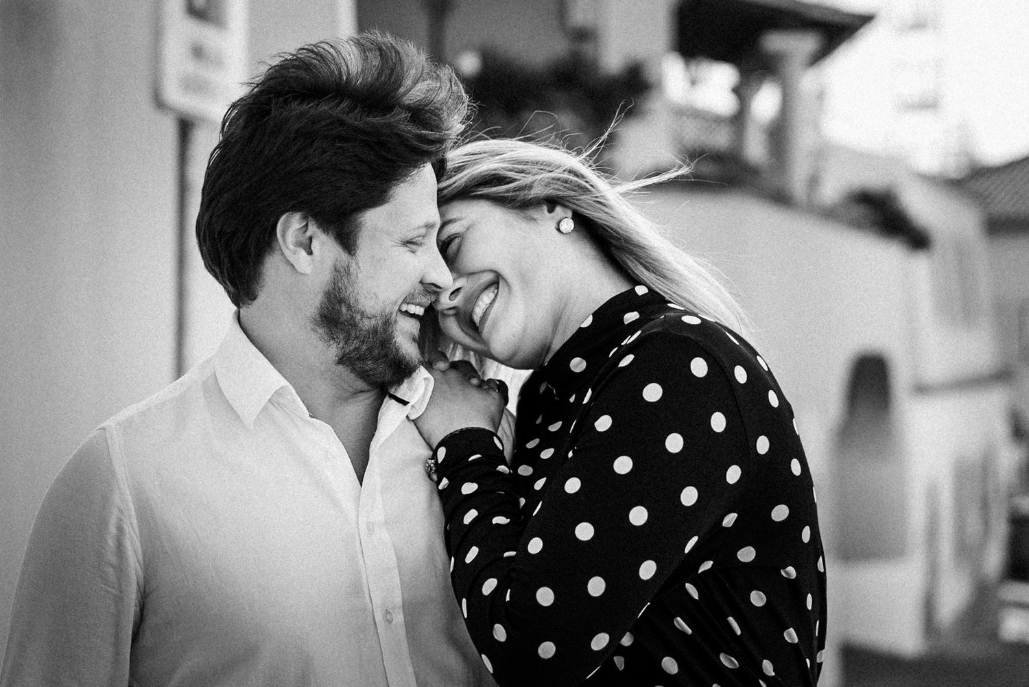A black and white photo shows a couple sharing an intimate moment while laughing together outdoors.