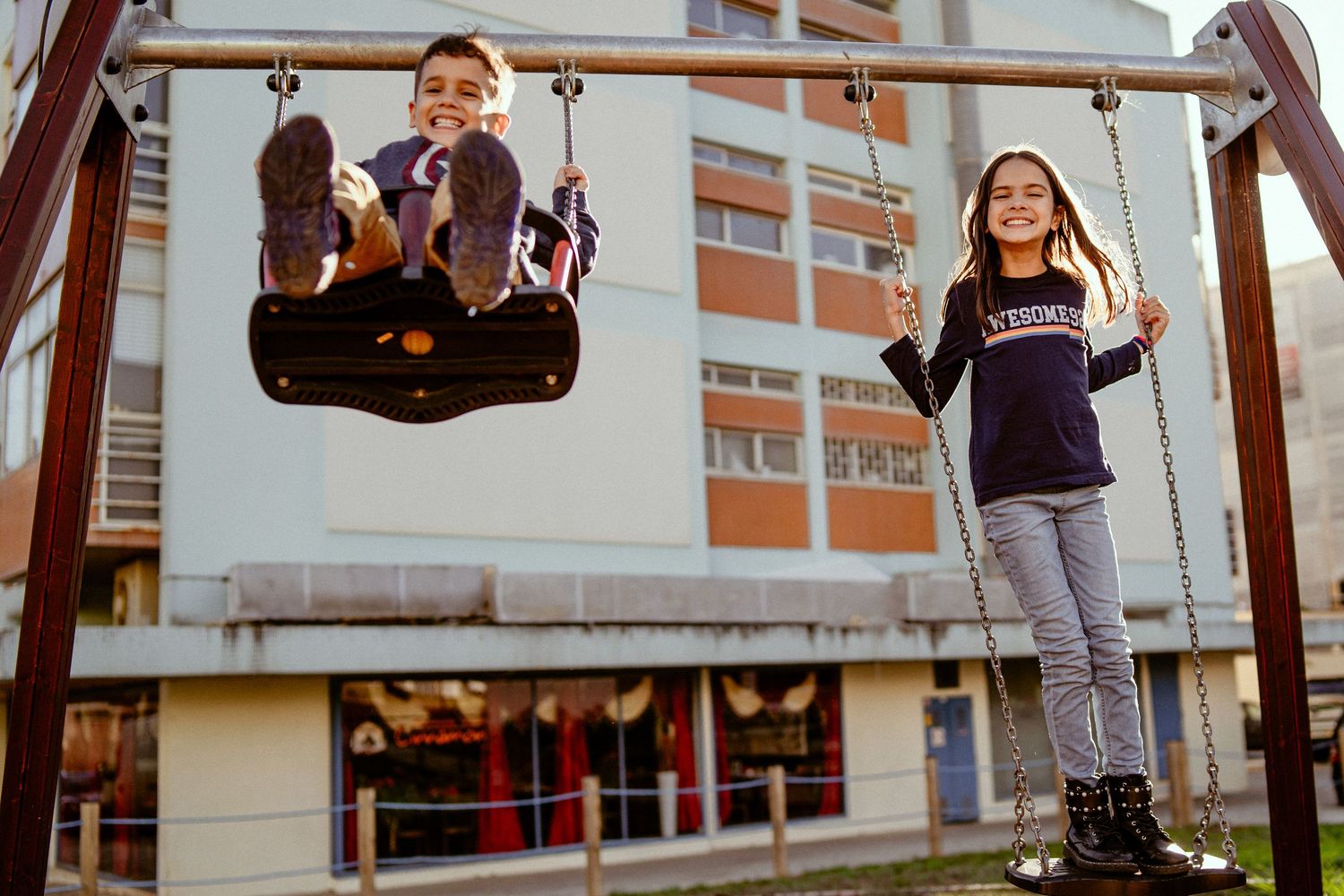 A smiling person sits on a playground swing wearing casual clothing at a park in daylight.