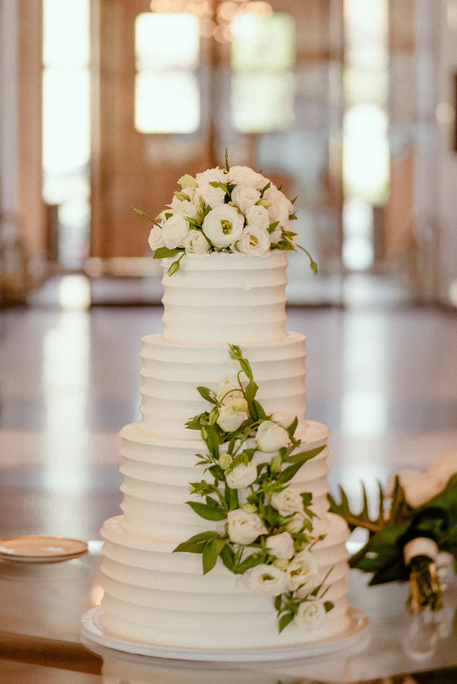 Three elegant white wedding cakes decorated with fresh white flowers and greenery sit on display tables in a light-filled venue.
