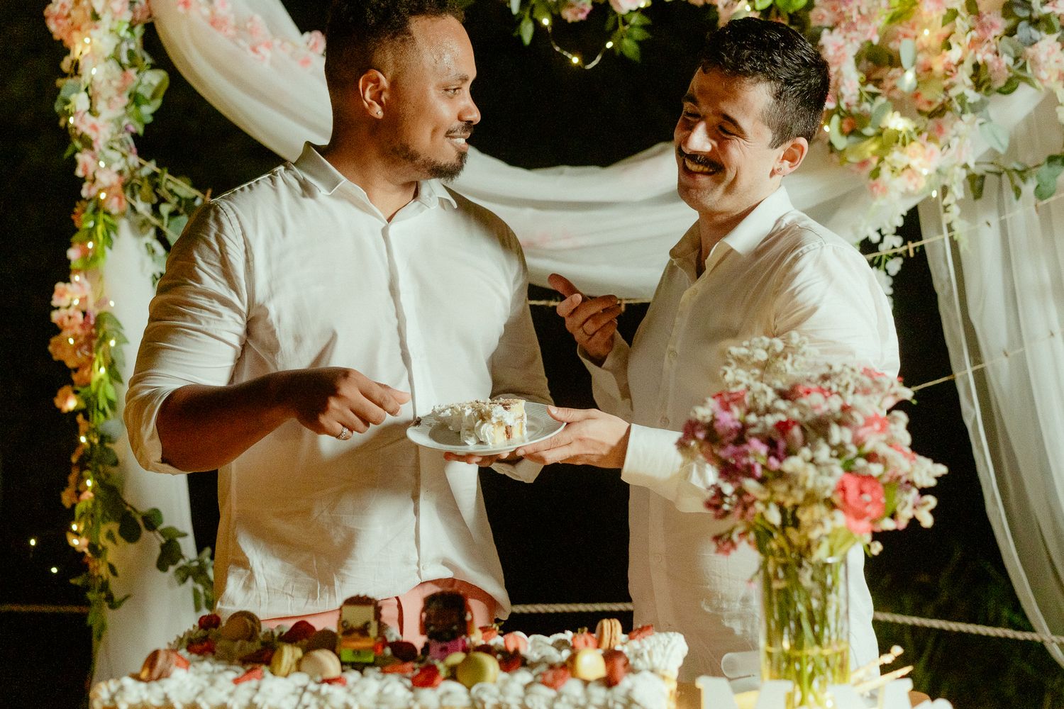 Two men in white shirts cutting a wedding cake together under floral arch decorations.