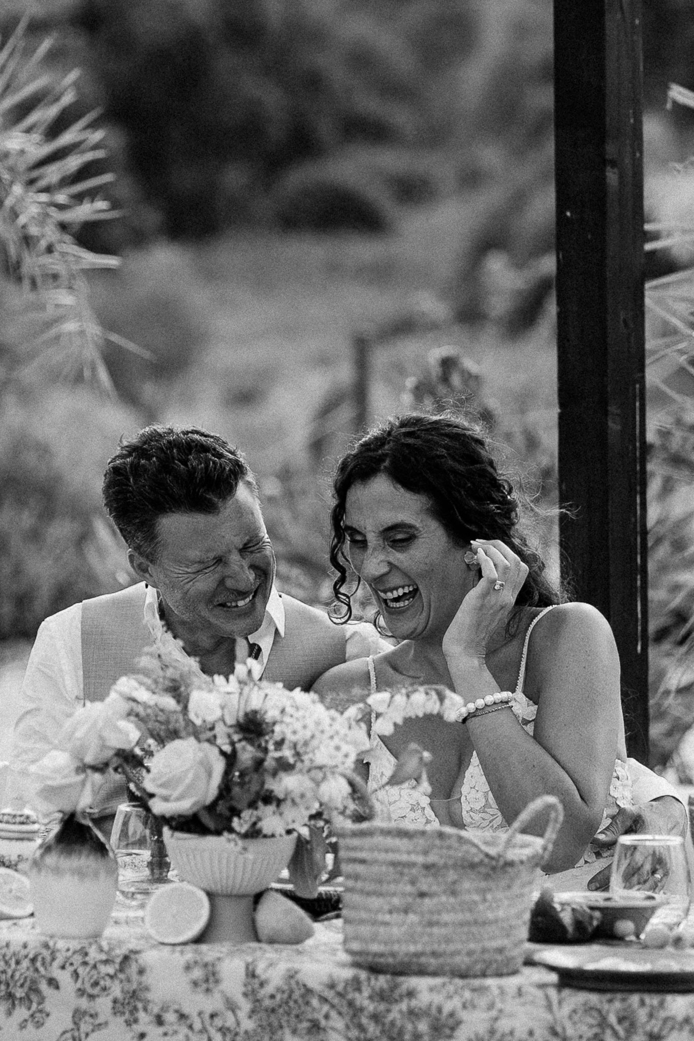 Black and white photo of a laughing couple at an outdoor wedding reception table with floral centerpiece.