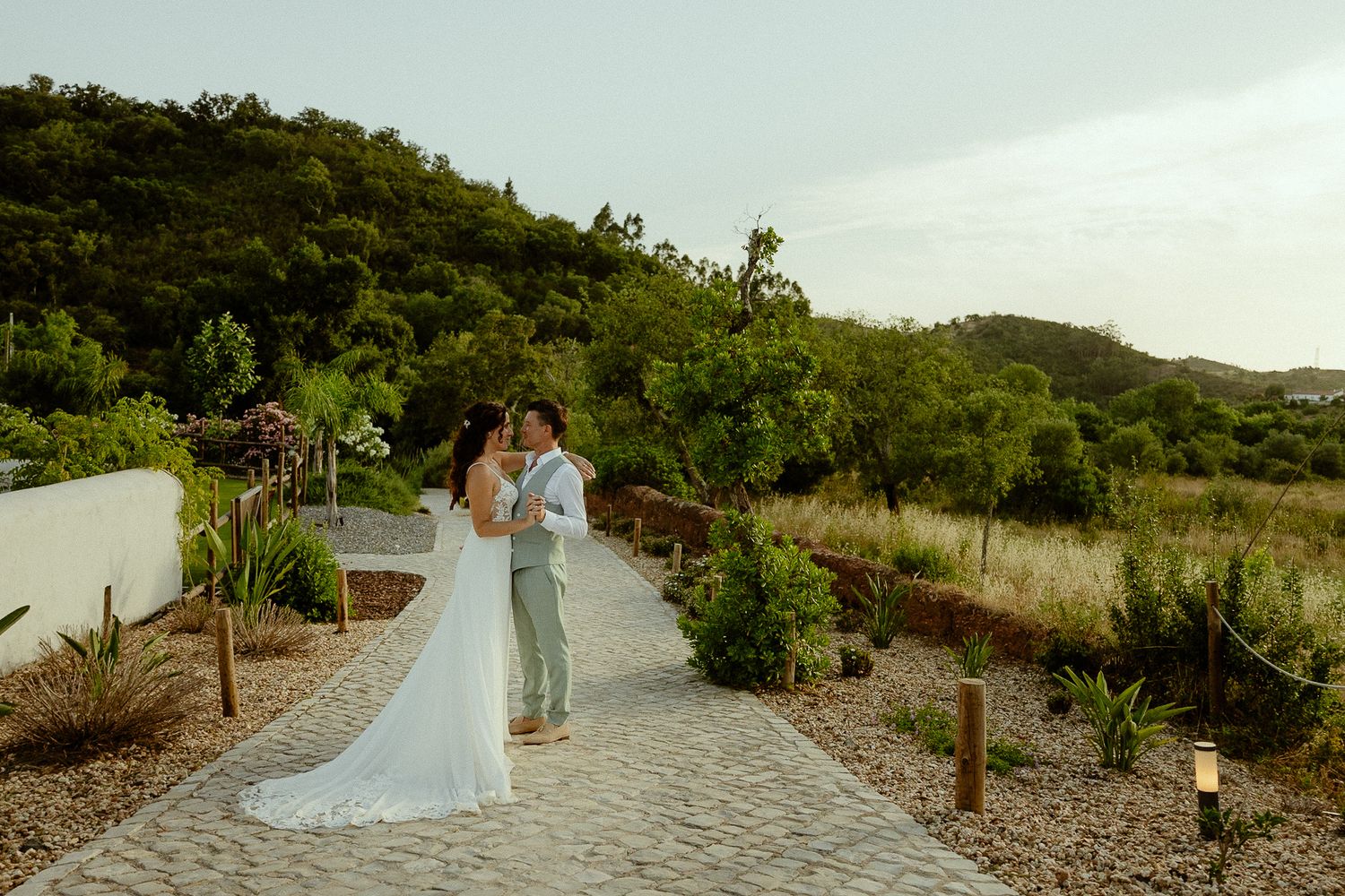 Bride and groom share a romantic moment on a stone path surrounded by lush greenery at an outdoor wedding venue.