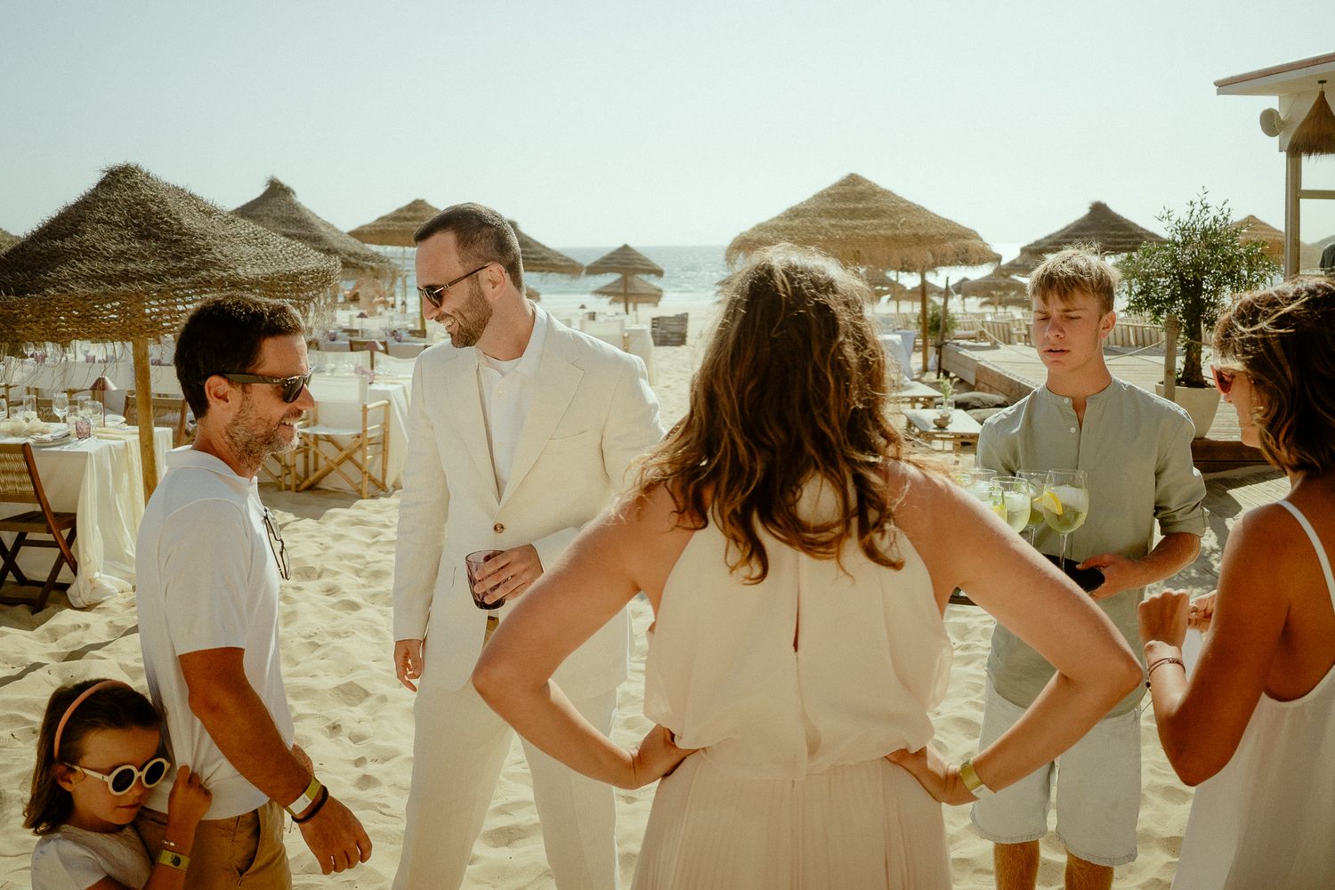 Group of elegantly dressed people socializing on a sunny beach with thatched umbrellas at a seaside event.