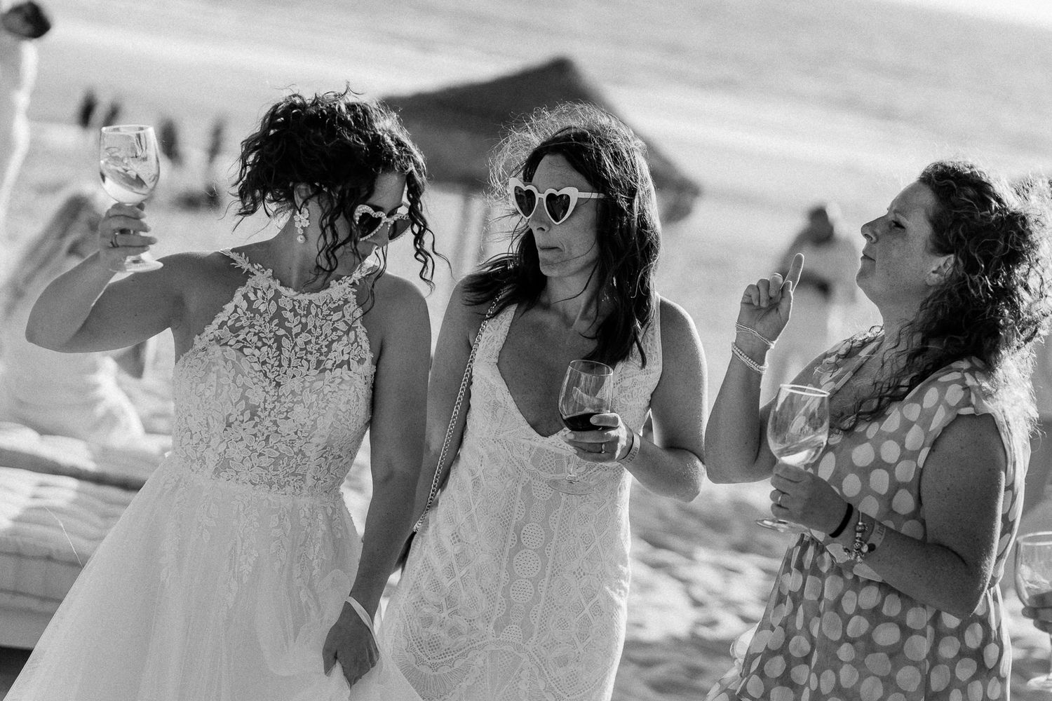 Two brides in white dresses celebrate with a friend on a beach, raising glasses in a joyful toast.