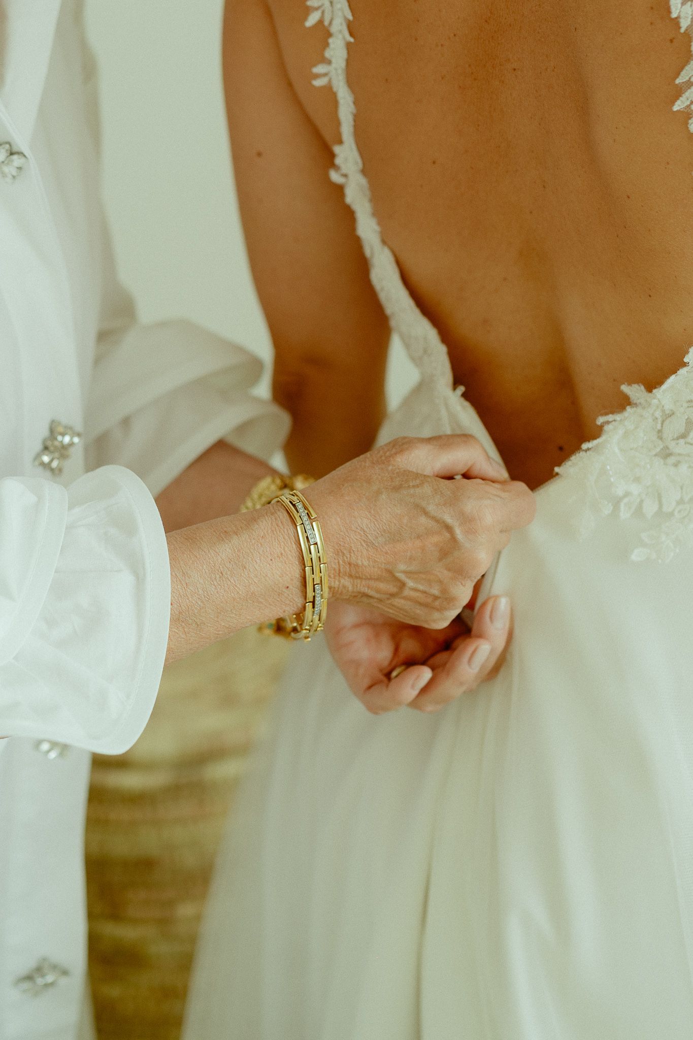 Older woman with gold bracelets fastening the back of a bride's open-back lace wedding dress.