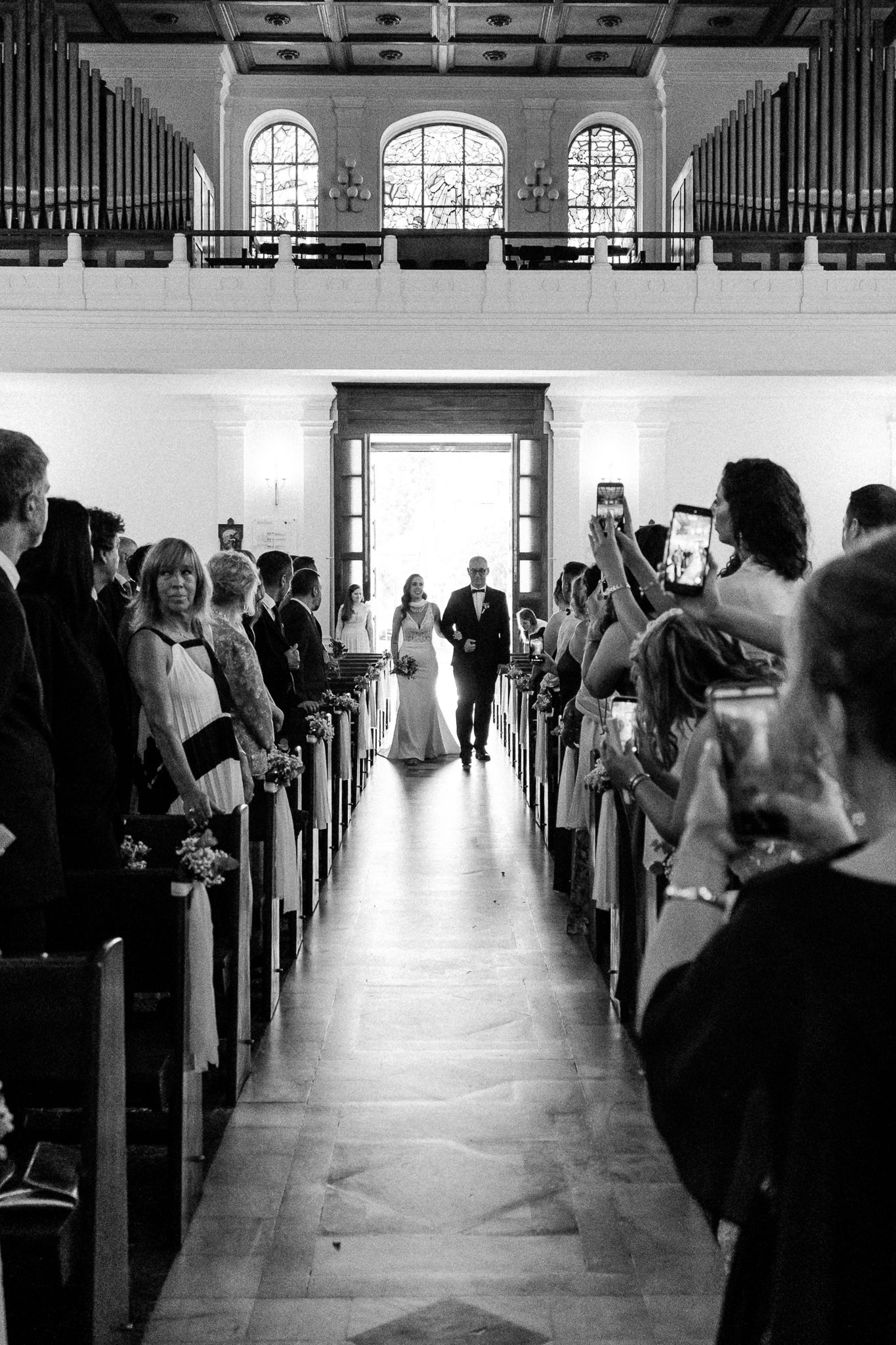 Black and white photo of a bride walking down the church aisle with guests watching during a wedding ceremony.
