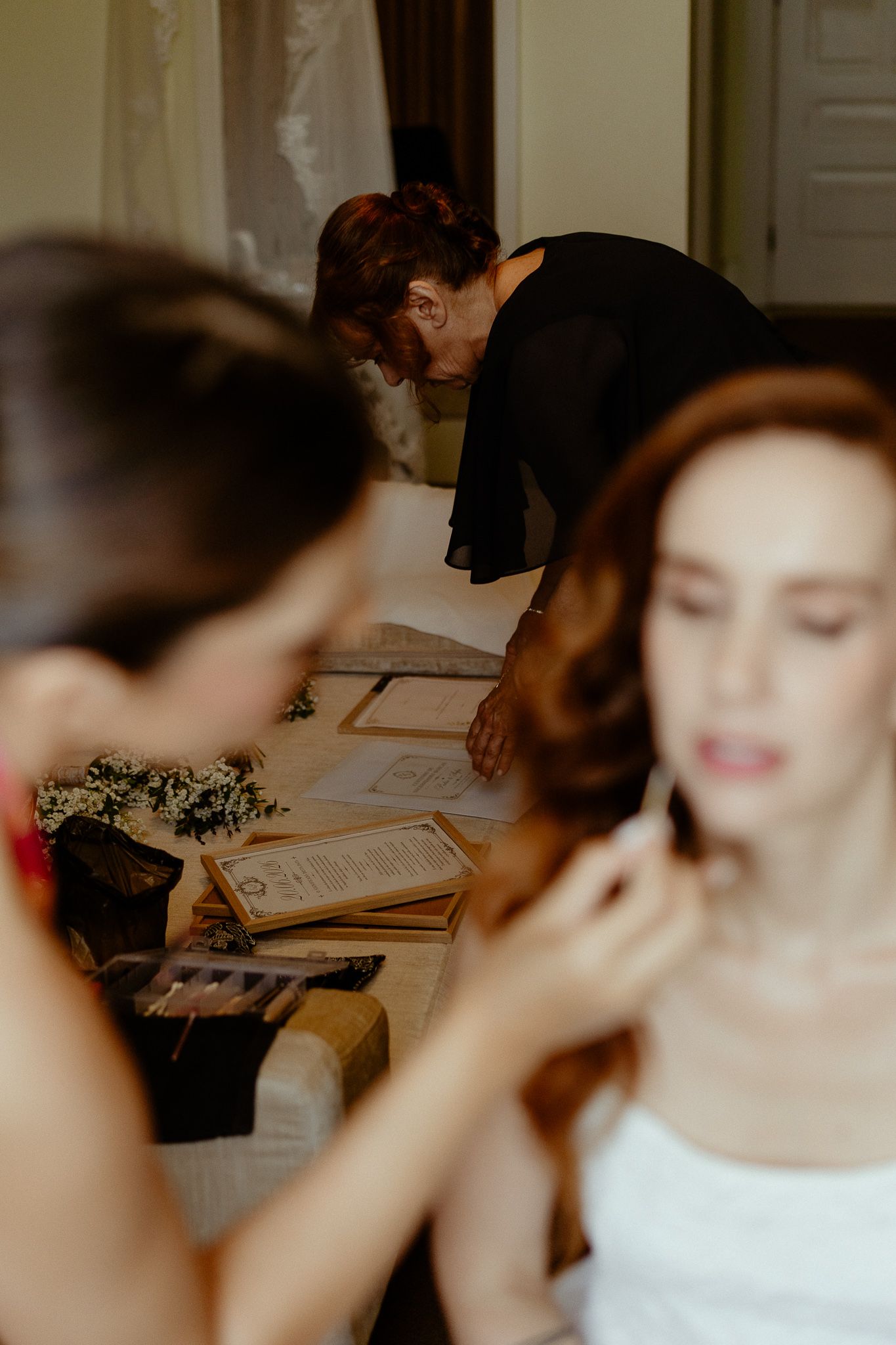 Bride getting ready with makeup artist while woman reviews wedding documents at a decorated table.