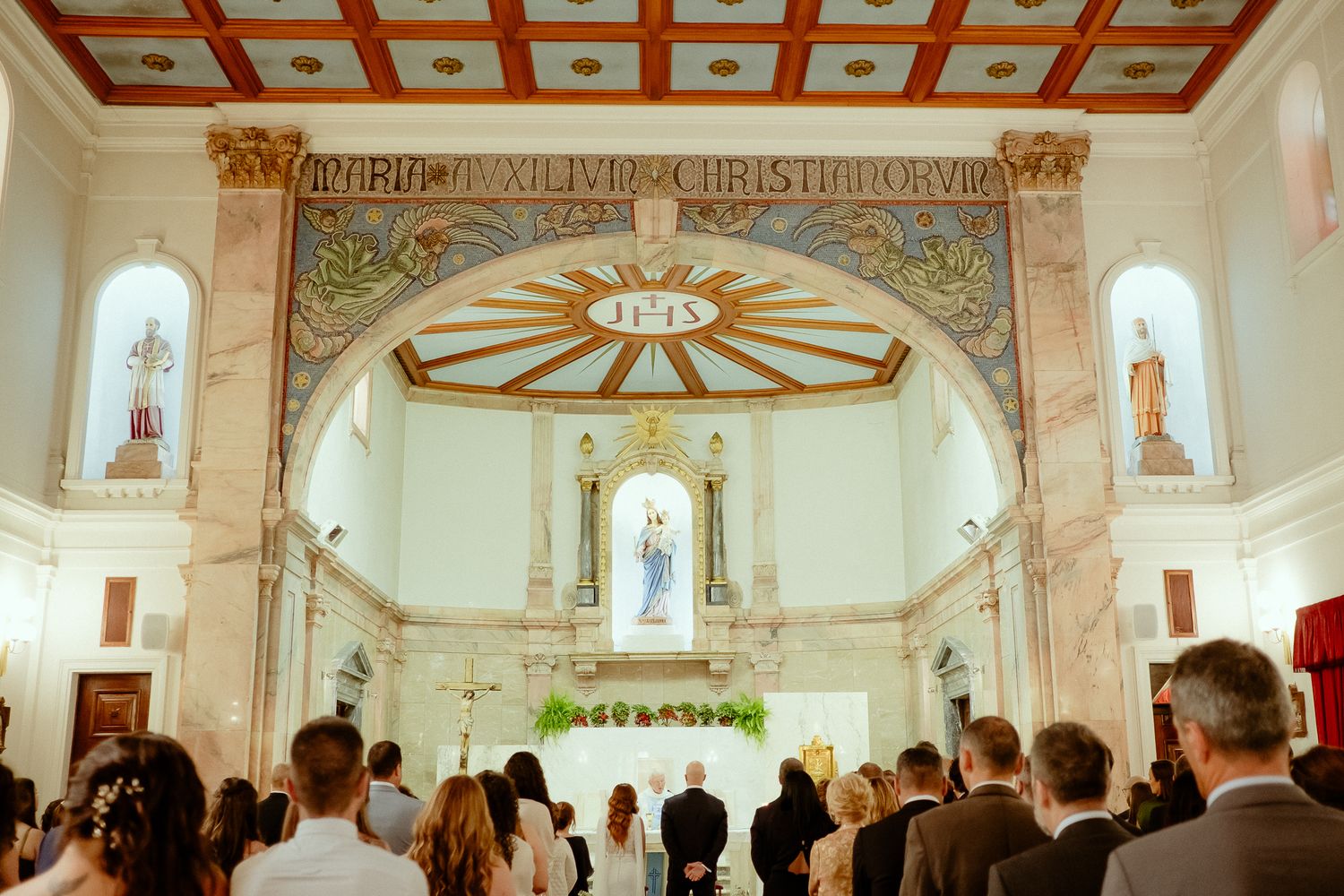 Elegant church wedding ceremony with guests standing before ornate altar featuring religious statue and decorative arch.