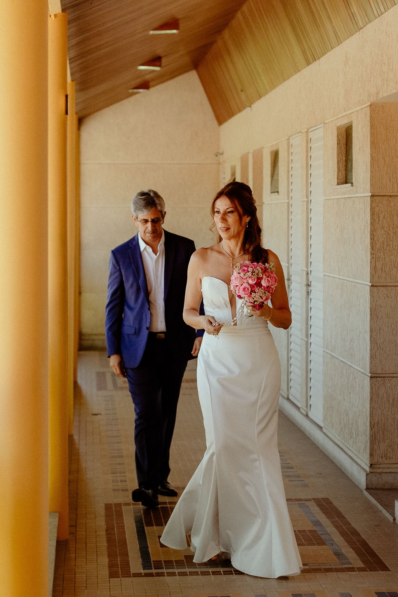 Wedding couple walks through a columned hallway with warm lighting and elegant architecture.
