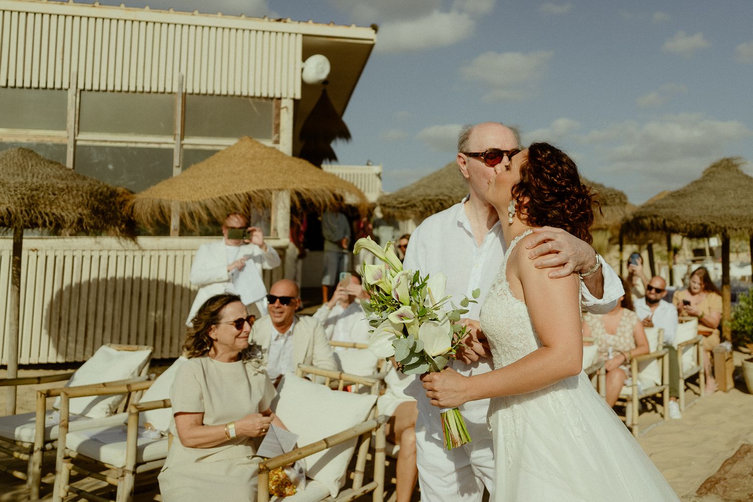 Bride and groom share a kiss at a sunny beach wedding ceremony with guests seated among thatched umbrellas.