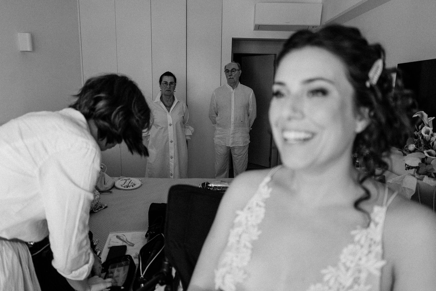 Black and white wedding photo of a smiling bride in lace dress with groom and guests in background.