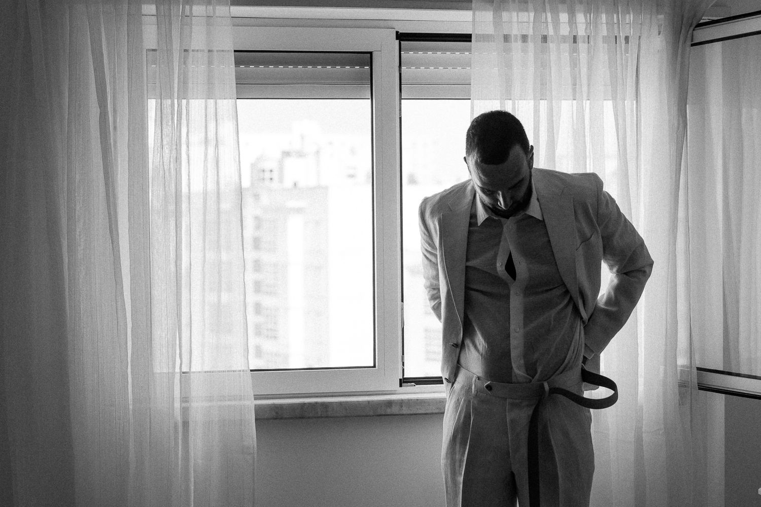 Black and white photo of a man in a suit adjusting his belt near a window with sheer curtains.