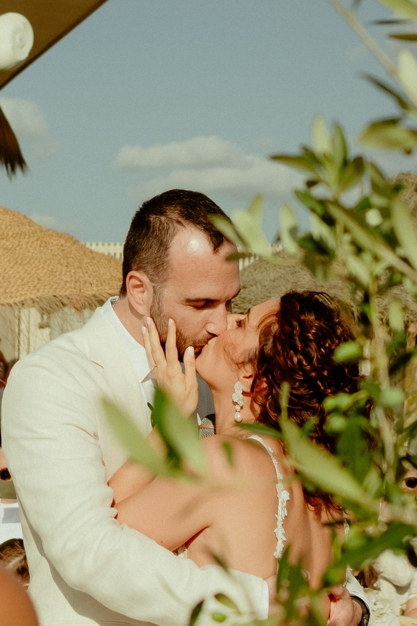 Bride and groom share their first kiss at an outdoor wedding ceremony surrounded by lush greenery under a blue sky.