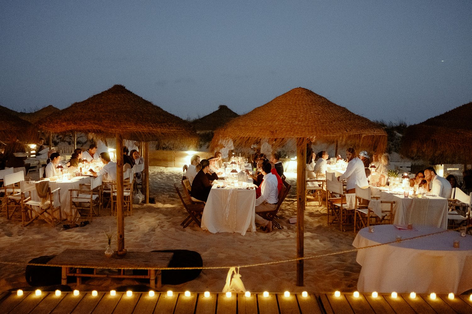 Romantic candlelit beach dinner reception at dusk with thatched umbrellas and elegant white tables.