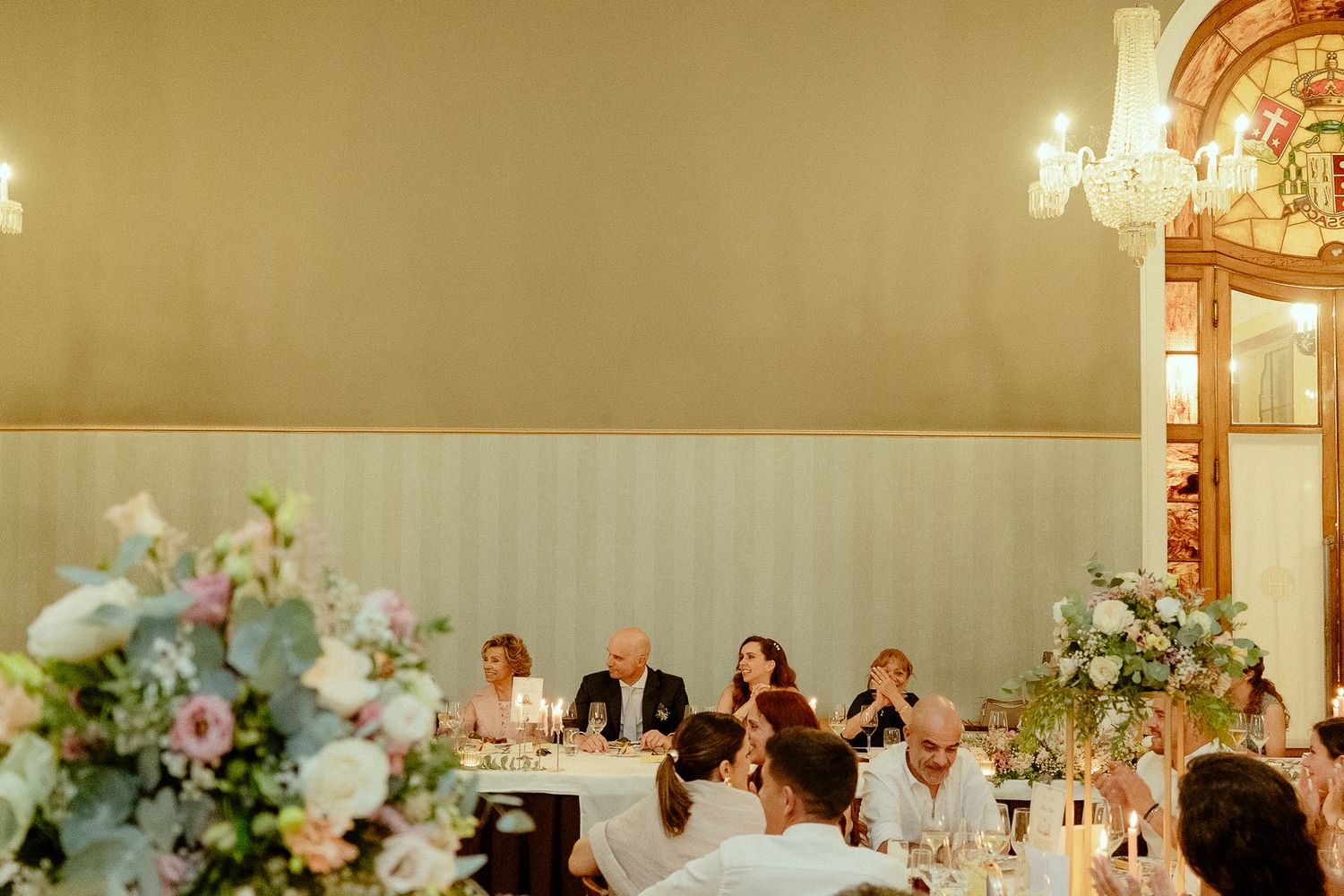 Elegant wedding reception in ornate ballroom with guests seated at decorated tables adorned with pastel floral arrangements.