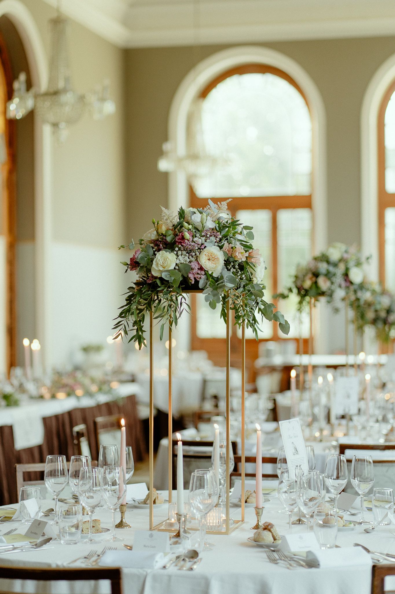 Elegant wedding reception hall with tall gold floral centerpieces, white tablecloths, candles, and arched windows.
