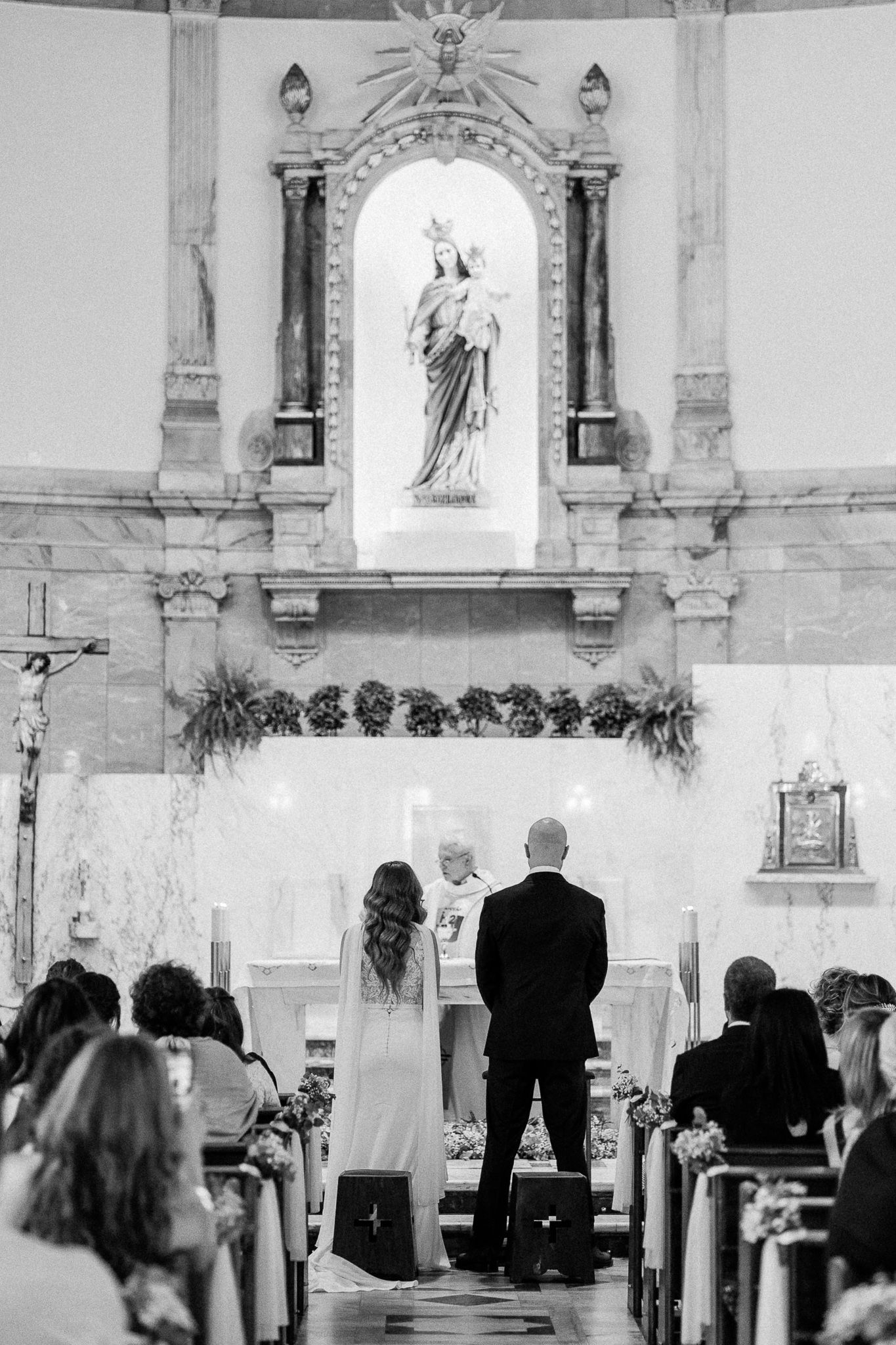 Black and white photo of a couple exchanging vows at a Catholic church altar with a Virgin Mary statue.