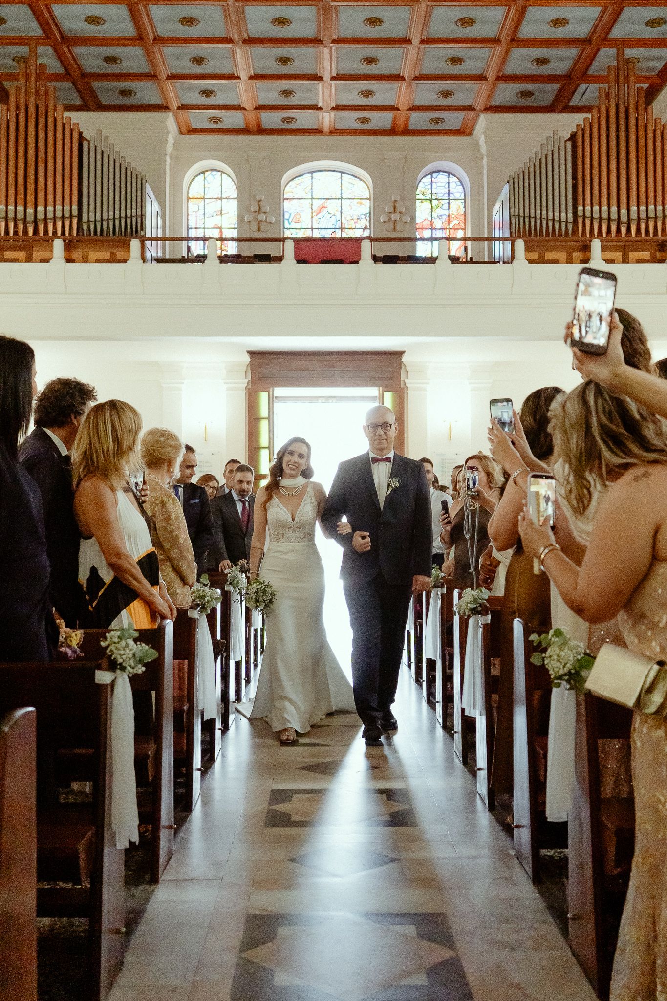 Bride walks down church aisle with father, guests photographing ceremony, organ pipes and stained glass windows visible.