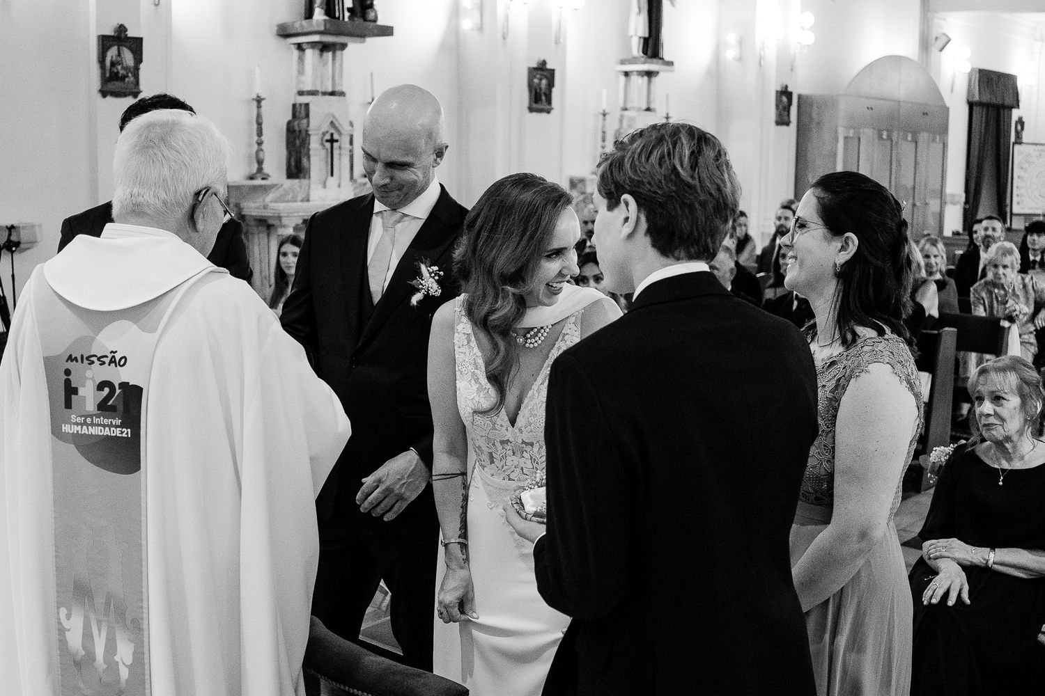 Black and white wedding ceremony photo inside a church with bride, groom, priest, and guests gathered together.