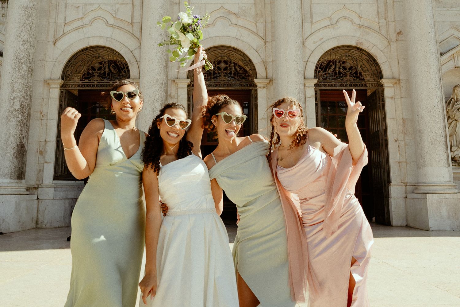 Four bridesmaids in pastel dresses celebrate outside ornate building, one holding bouquet aloft.
