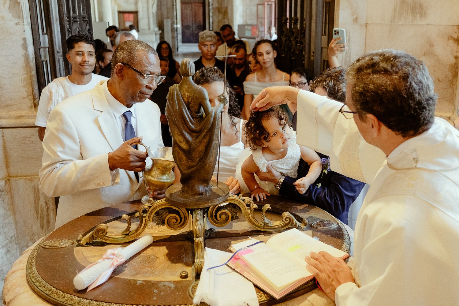 A priest performs a baptism ceremony, gently pouring water over a young child's head at a church font.