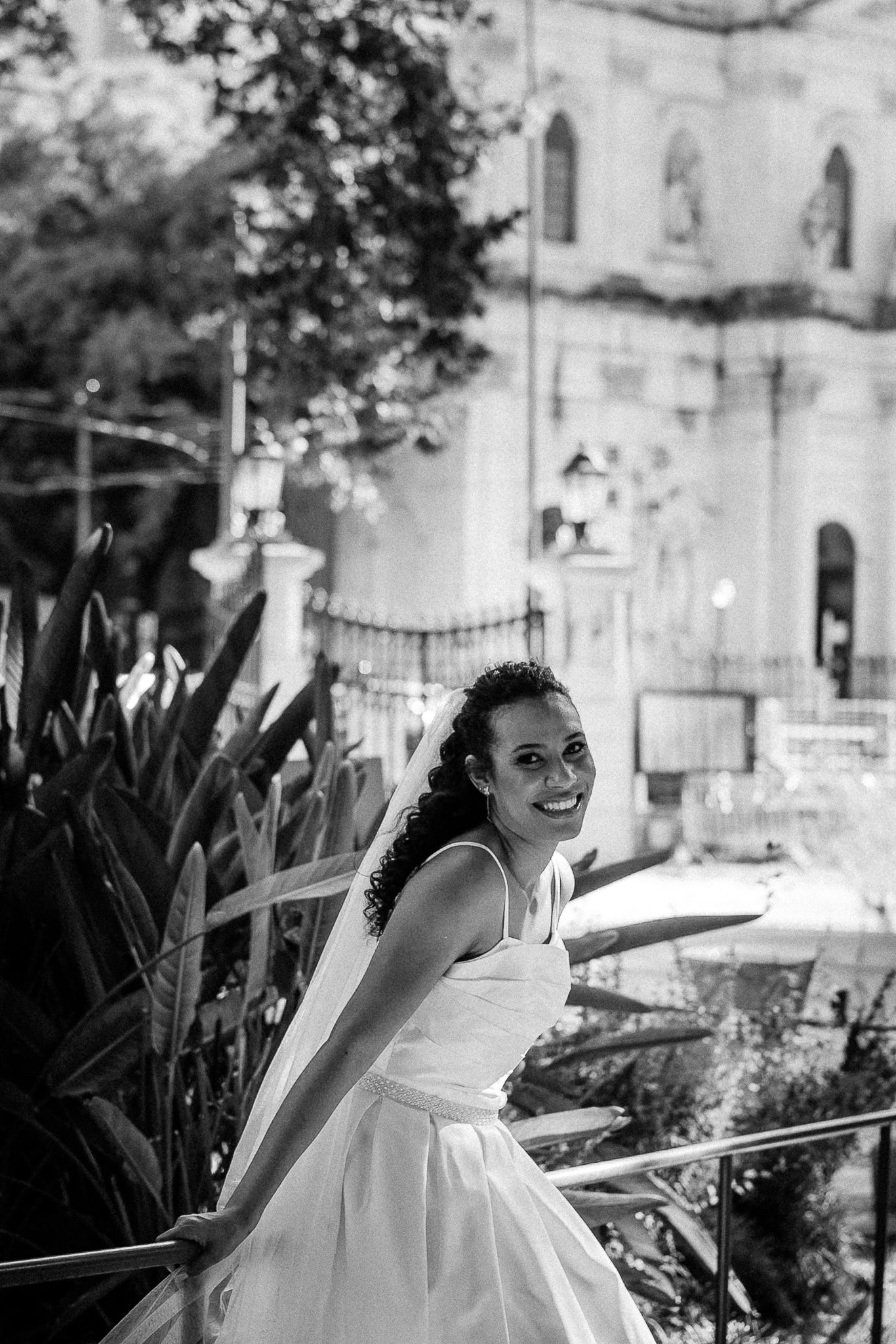 Black and white photo of a smiling bride in white gown leaning on railing with blurred church background.