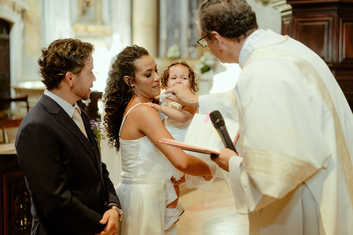 Bride and groom with priest during wedding ceremony in church, bride holding a young child in white dress.