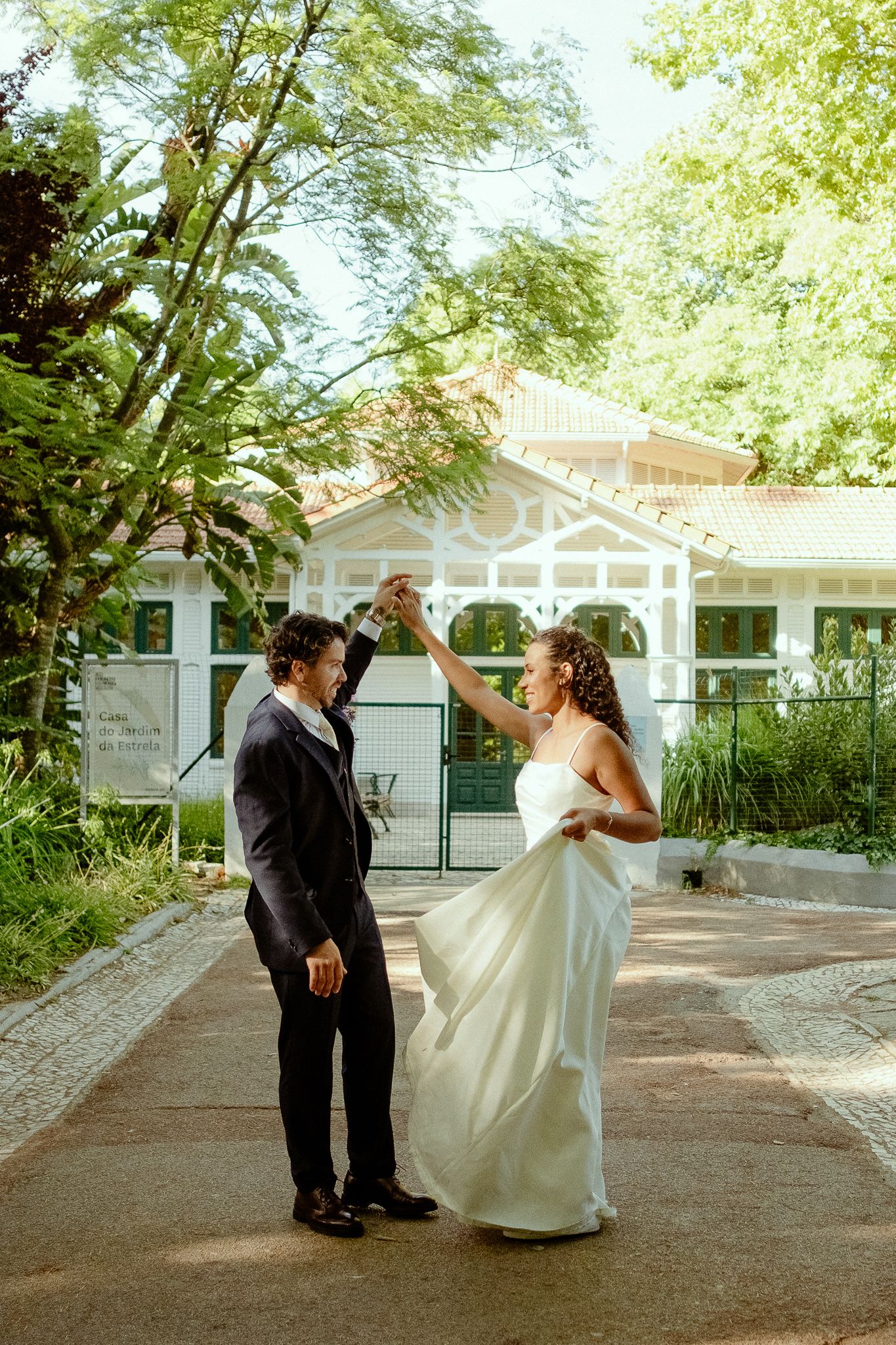A bride and groom dance together outdoors in a lush garden setting, with a white pavilion building in the background.
