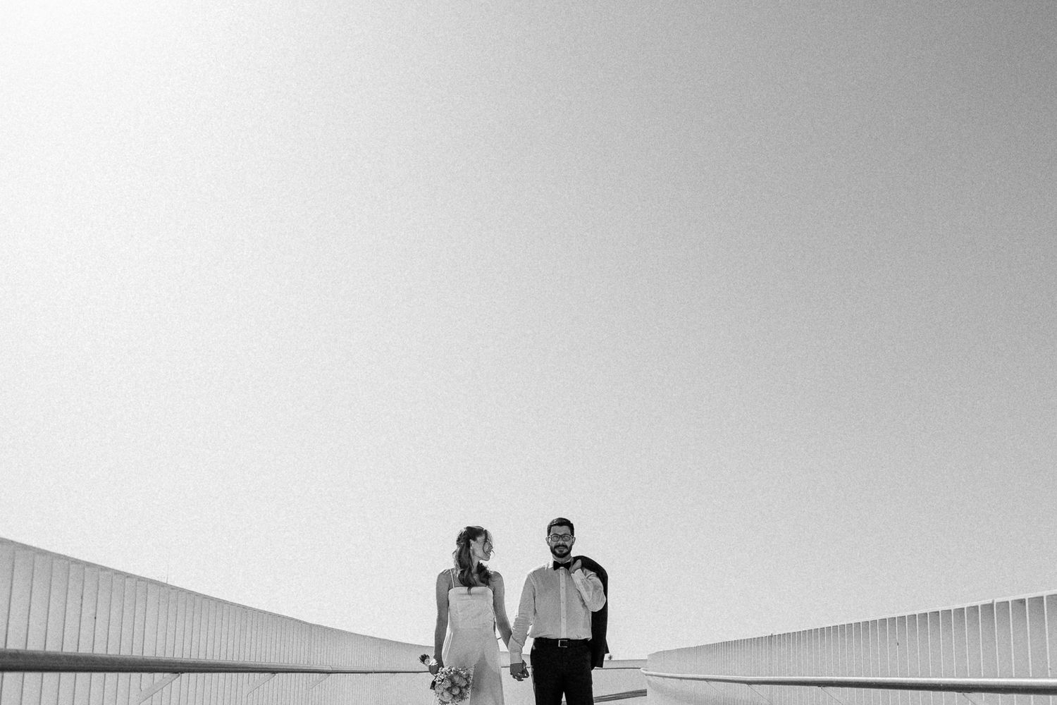 Black and white photo of a bride and groom holding hands outdoors under a clear sky on their wedding day.